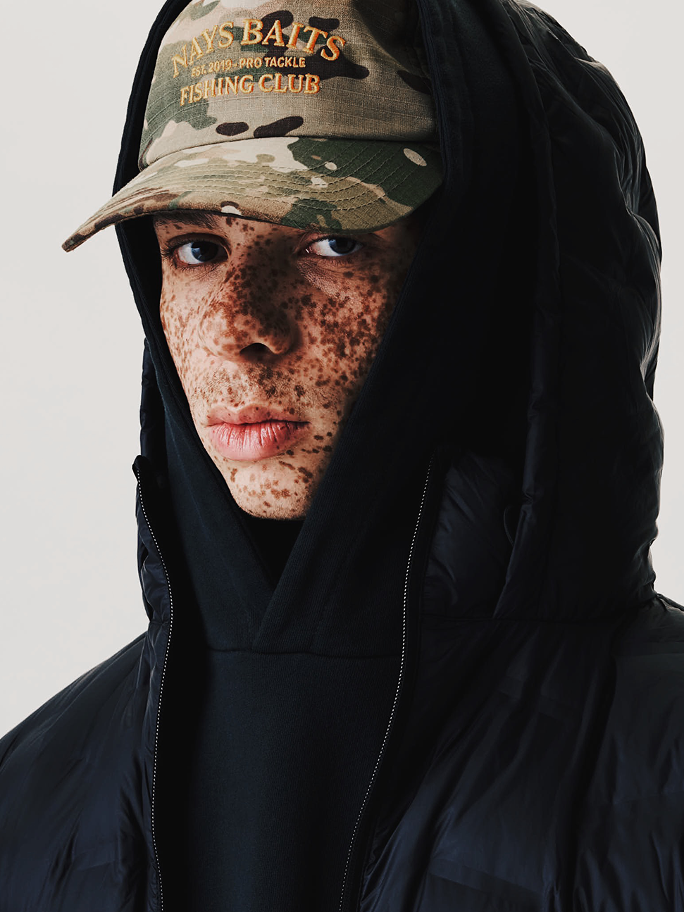 Model mit camouflagefarbener NAYS Fishing Club Cap, schwarzem Hoodie und schwarzer Pufferjacke vor cleanem Studiohintergrund.