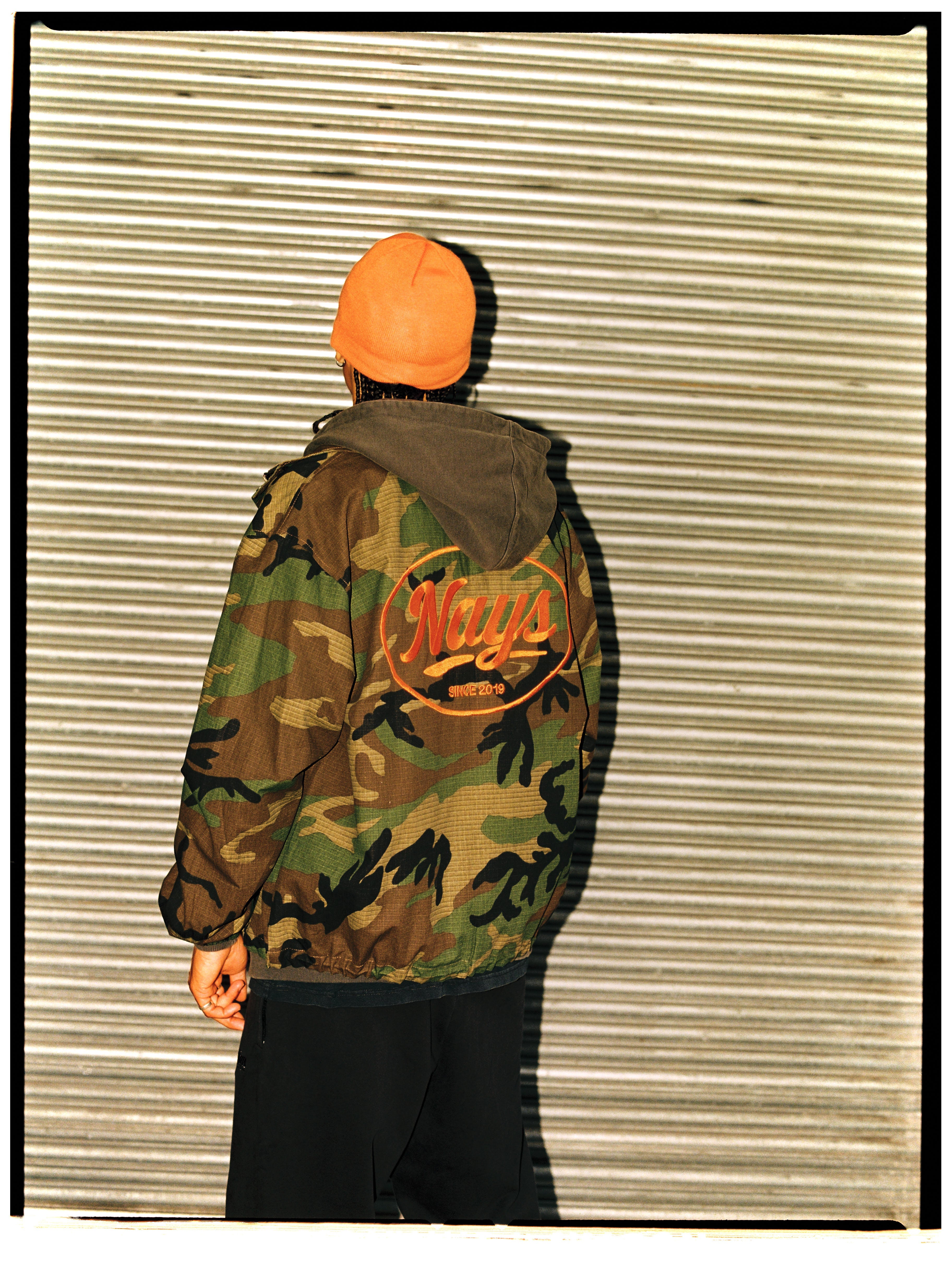 Mann von hinten in Nays Camouflage-Jacke mit Logo-Stickerei und orangefarbener Beanie.
