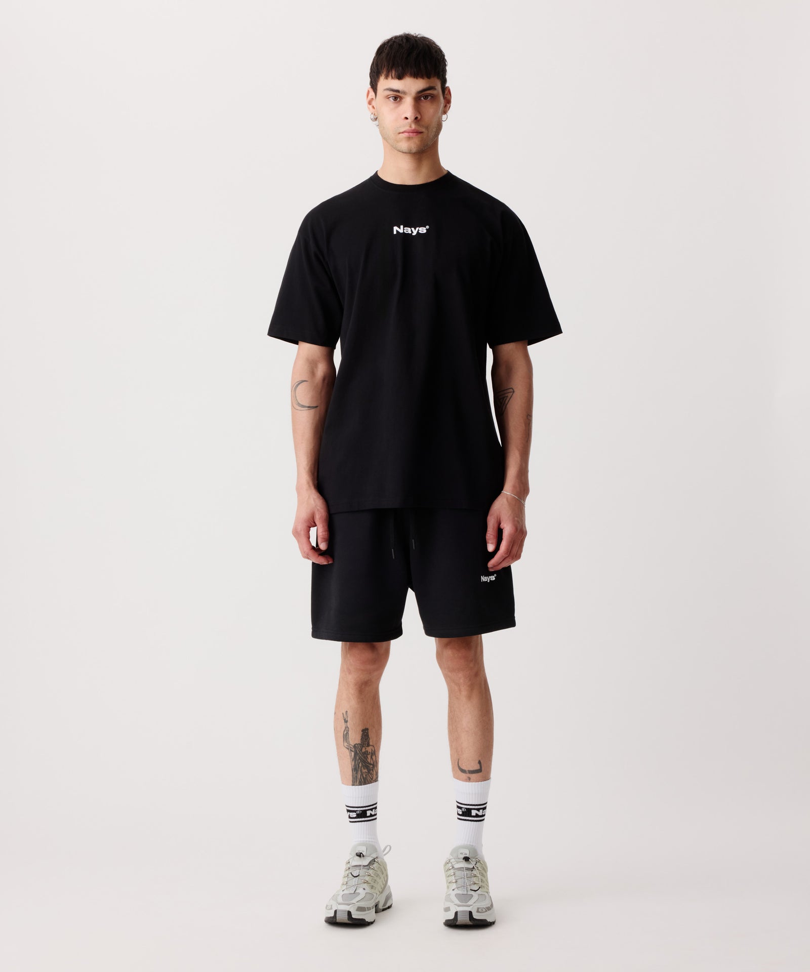 Das Bild zeigt das schwarze Basic T-Shirt mit Front-Print getragen von einem Mann. Dazu trägt er eine kurze, schwarze Hose, weiße Logo-Socken und helle Turnschuhe.