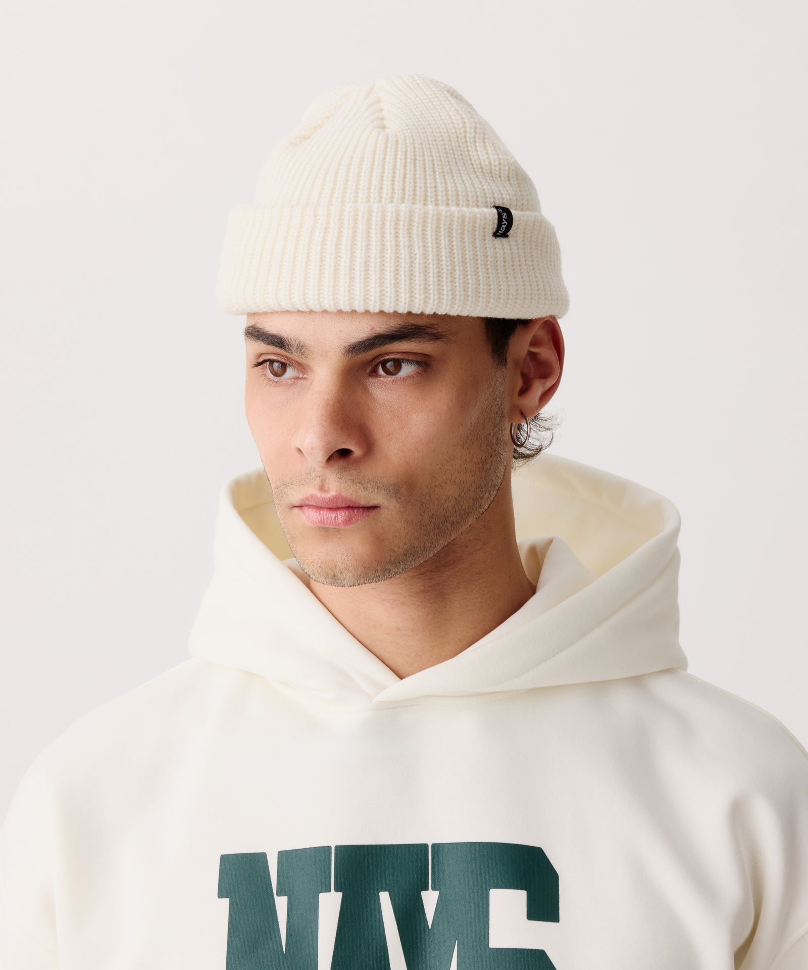 Creme-farbige Strickmütze mit gerippter Textur getragen von einem Mann. Er schaut zur Seite und trägt einen farblich passenden Pullover mit Kapuze.