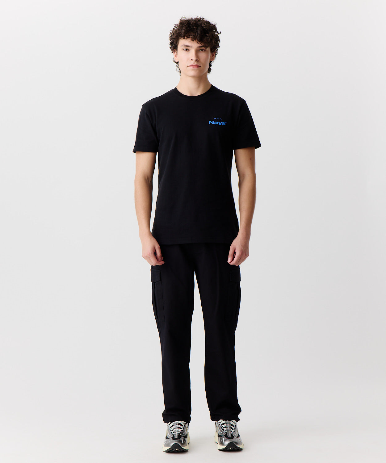 Das Bild zeigt die Nays Hose aus Baumwolle getragen von einem Mann. Er trägt ein schwarzes T-Shirt mit blauem Logo und helle Turnschuhe dazu.