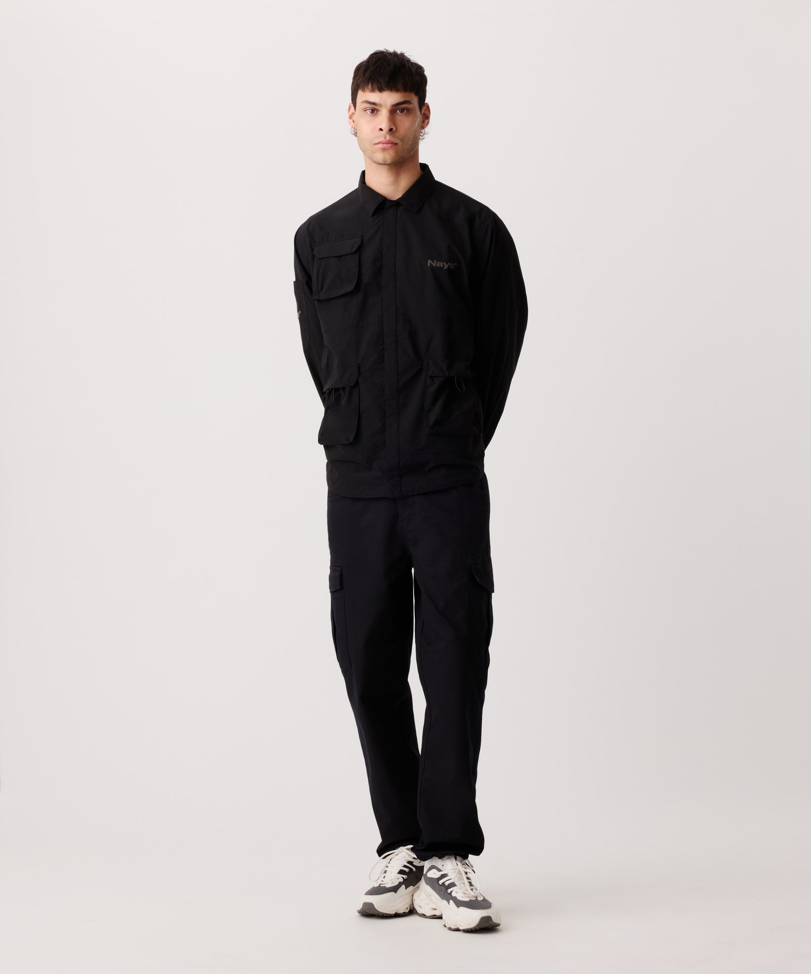 Nays Utility Shirt, eine leichte Shirt-Jacke mit Reverskragen, aufgesetzten Taschen, wasserabweisendem Finish und elastischen Drawstrings getragen von einem Mann in der frontalen Ansicht. Er trägt dazu eine schwarze Hose und helle Sneaker.