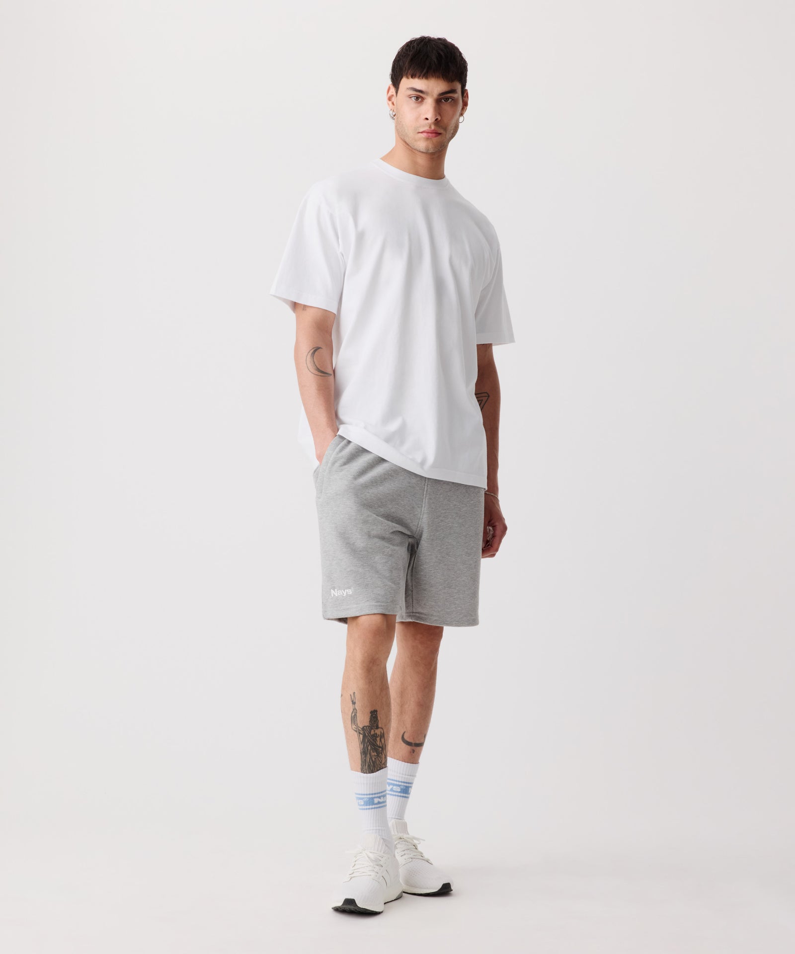 Das Bild zeigt die Nays Shorts in grau getragen von einem Mann. Er trägt ein weißes T-Shirt, weiße Logo Socken und helle Turnschuhe.