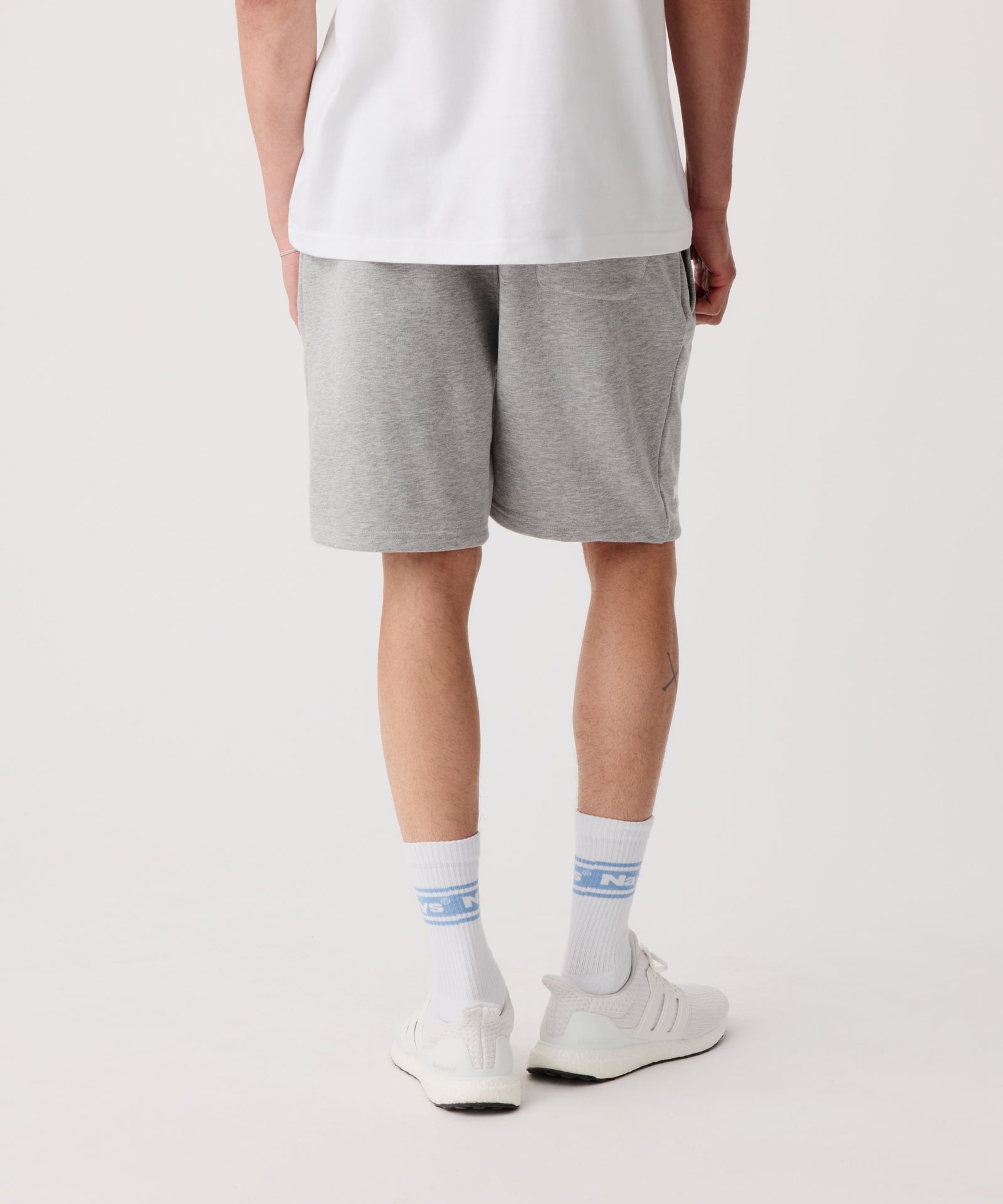 Das Bild zeigt die Nays Shorts in grau getragen von einem Mann in der Rückansicht. Er trägt ein weißes T-Shirt, weiße Logo Socken und helle Turnschuhe.
