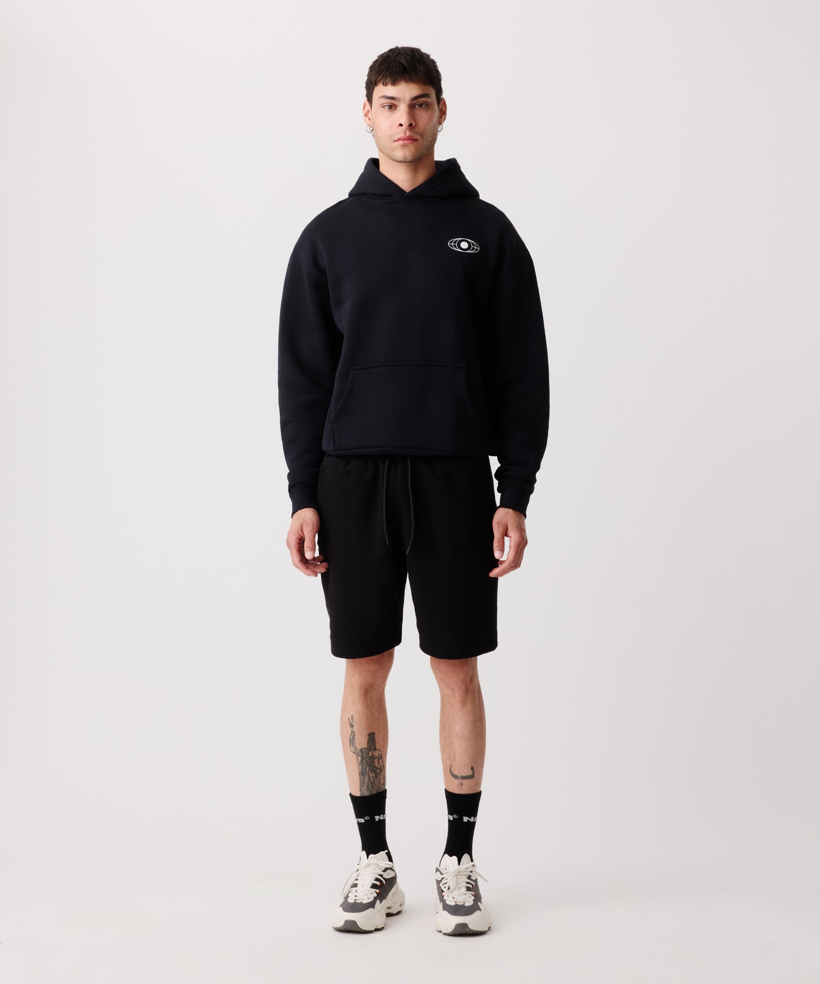 Das Bild zeigt die Nays Shorts in schwarz getragen von einem Mann. Er trägt einen farblich passenden Pullover mit Kapuze, schwarze Logo Socken und helle Turnschuhe.