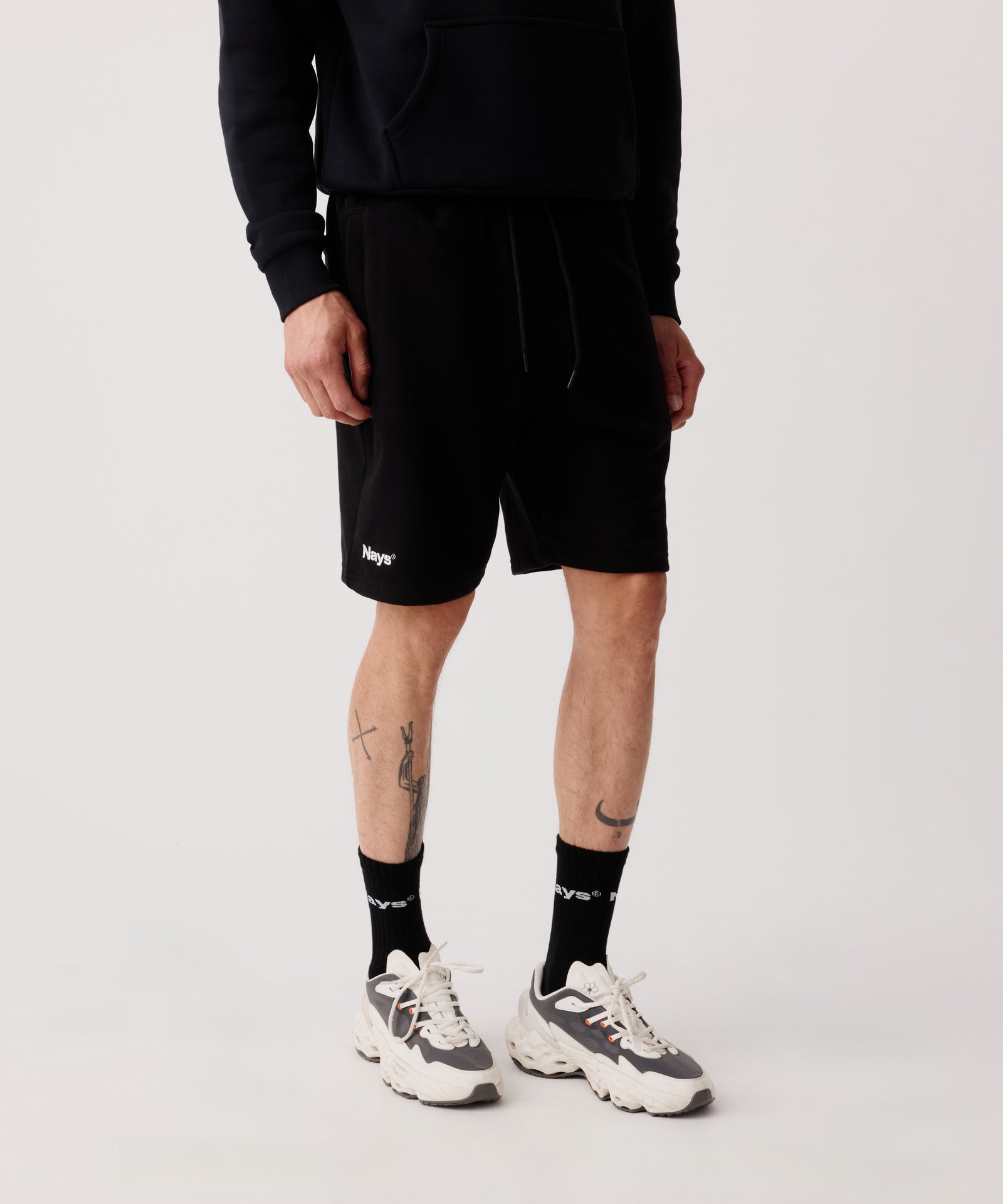 Das Bild zeigt die Nays Shorts in schwarz getragen von einem Mann. Er trägt einen farblich passenden Pullover, schwarze Logo Socken und helle Turnschuhe.
