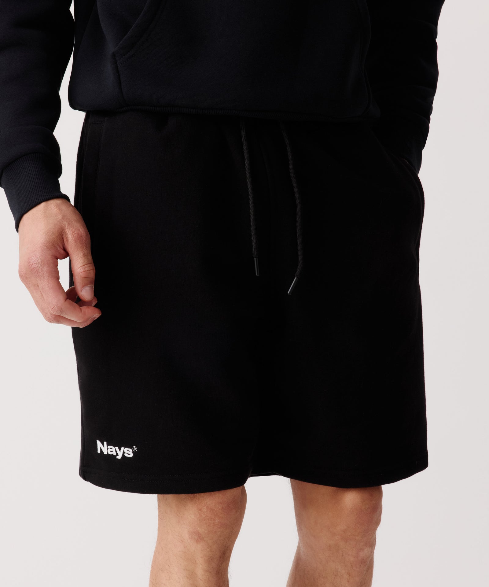 Der Bildausschnitt zeigt die Nays Shorts in schwarz getragen von einem Mann. Der Fokus liegt auf den Kordelzügen im Bund, eine Hand befindet sich in der Seitentasche.