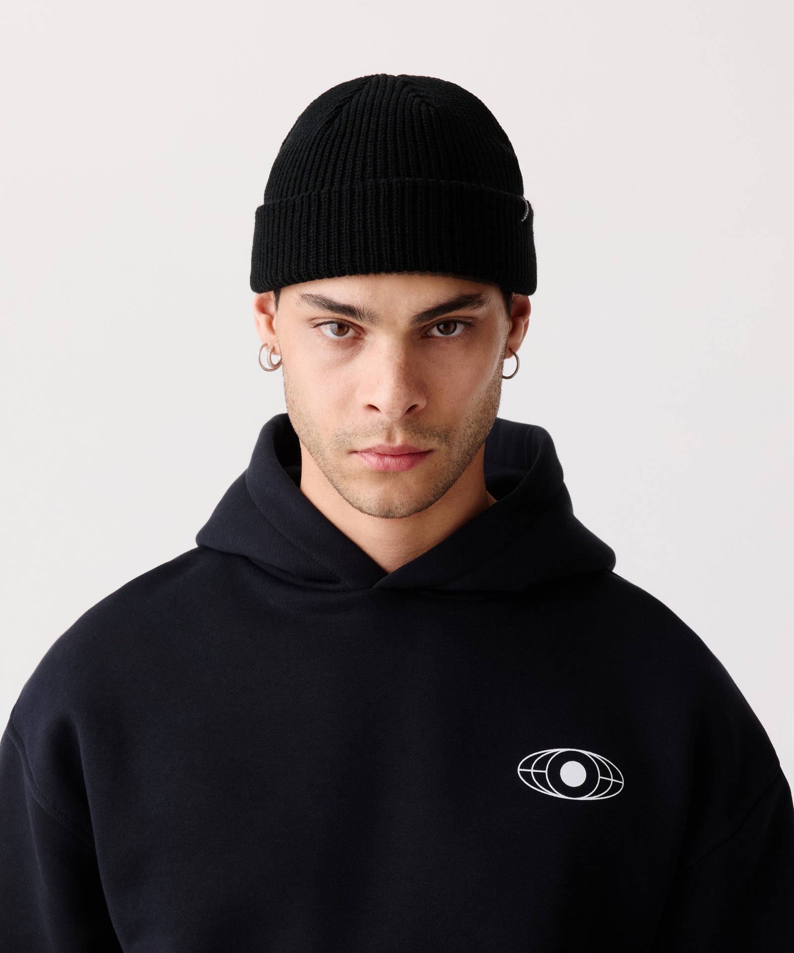 Schwarze Strickmütze mit gerippter Textur getragen von einem Mann. Dazu trägt er einen schwarzen Kapuzen-Pullover mit weißem Logo-Aufdruck.