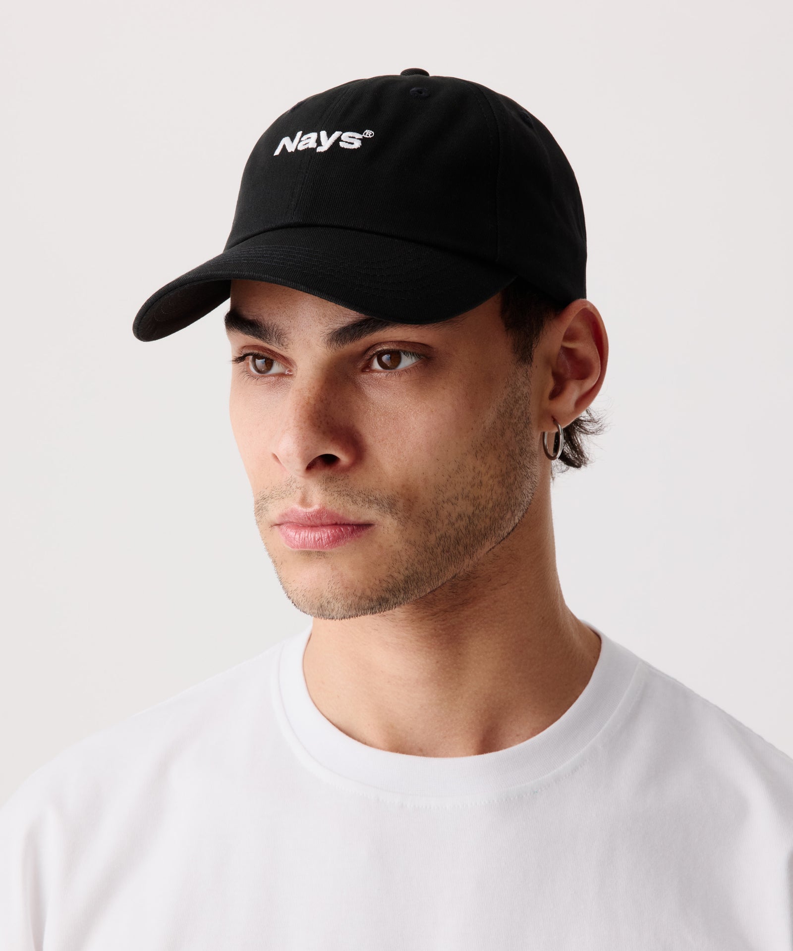 Eine sportliche Schirm-Mütze mit mittig gesticktem Nays Logo. Das Model trägt dazu ein weißes T-Shirt und schaut leicht schräg an der Kamera vorbei.