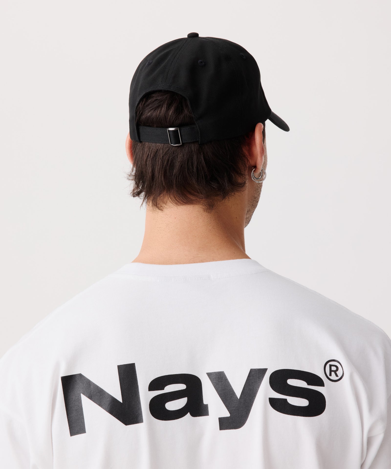 Eine sportliche Schirm-Mütze getragen von einem Mann. Gut zu erkennen ein größen-verstellbarer Riemen. Das Model trägt dazu ein weißes T-Shirt mit großem Nays Logo Print auf dem Rücken.