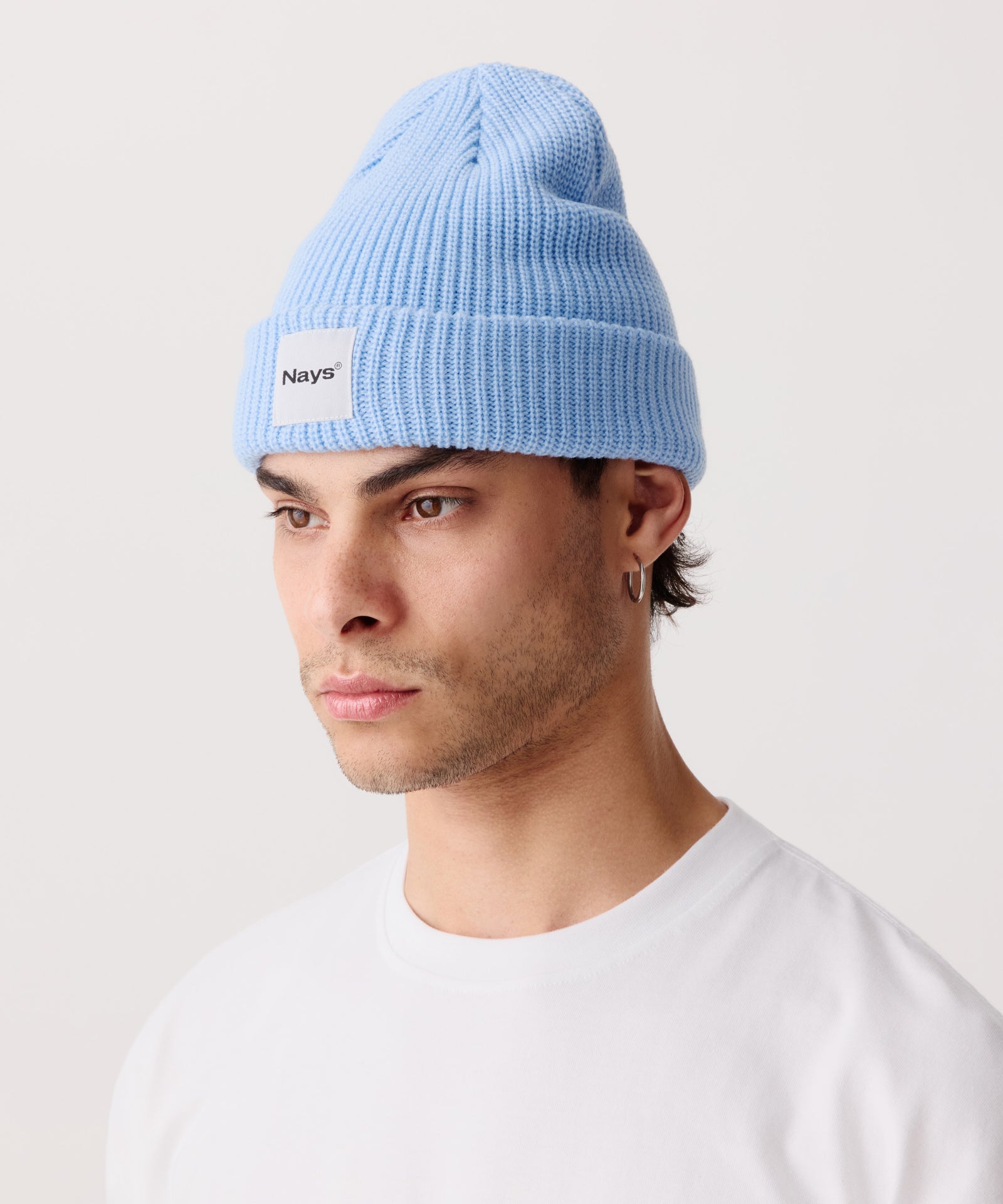 Hell-Blaue Strickmütze mit breitem Umschlag getragen von einem Mann. Mittig gut zu erkennen ein quadratischer Aufnäher mit mittig gesetztem Nays Logo.