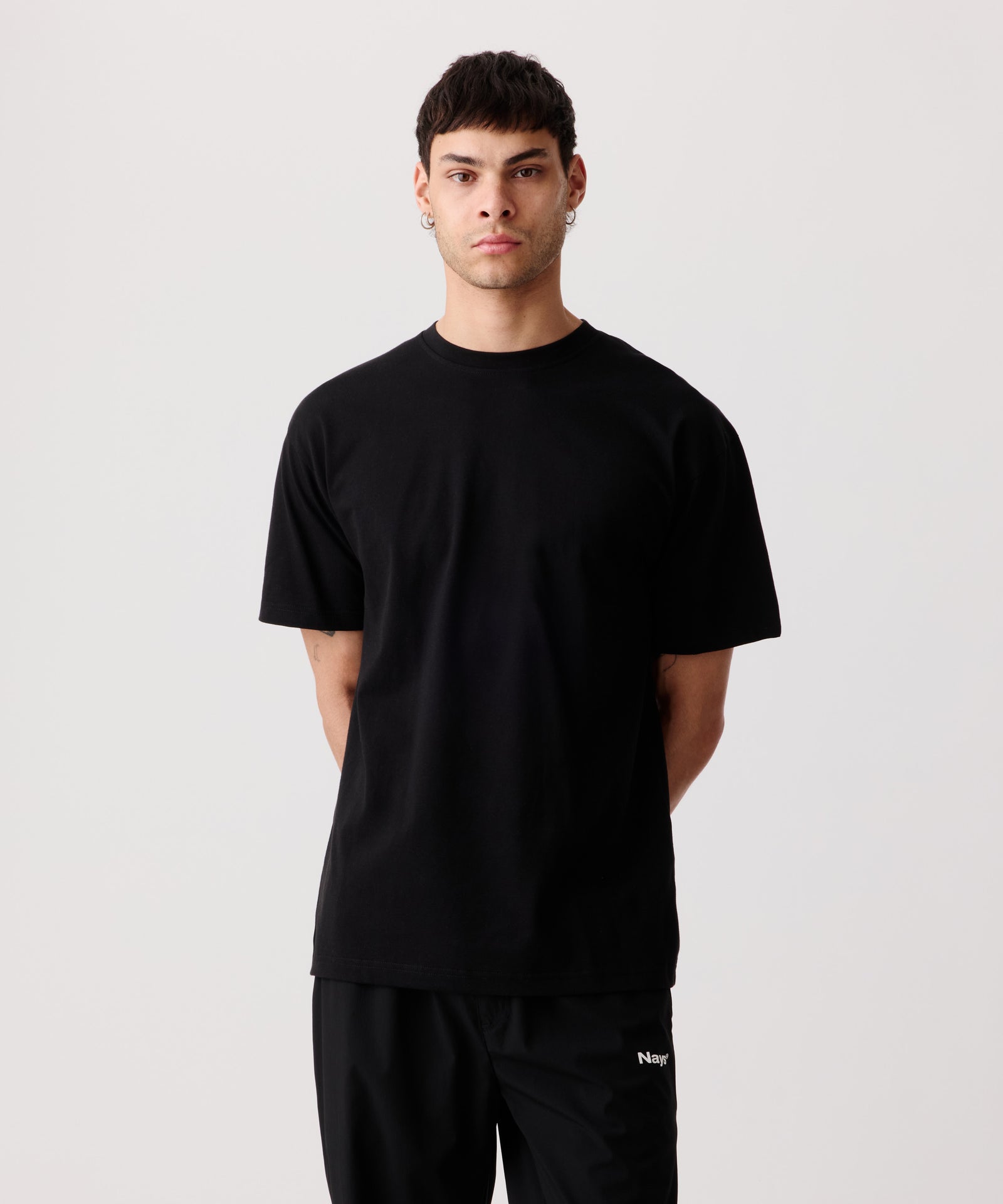 Ein Mann trägt das weiße Logo-T-Shirt mit verschränkten Armen auf dem Rücken. Dazu kombiniert er eine schwarze Hose mit weißem „Nays“-Logo-Schriftzug.