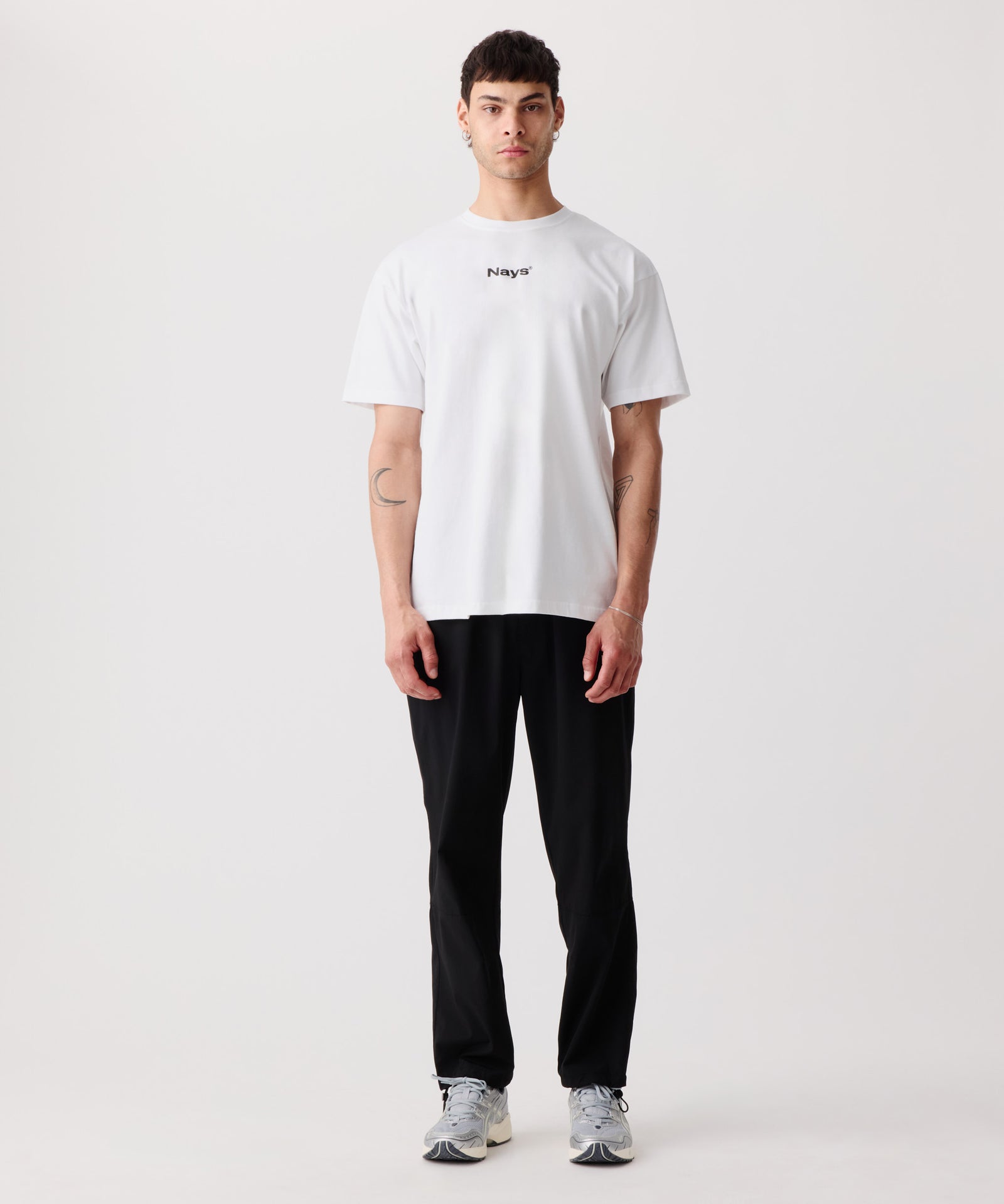 Das Bild zeigt das weiße Basic T-Shirt mit Front-Print getragen von einem Mann. Dazu trägt er eine lange, schwarze Hose und helle Turnschuhe.