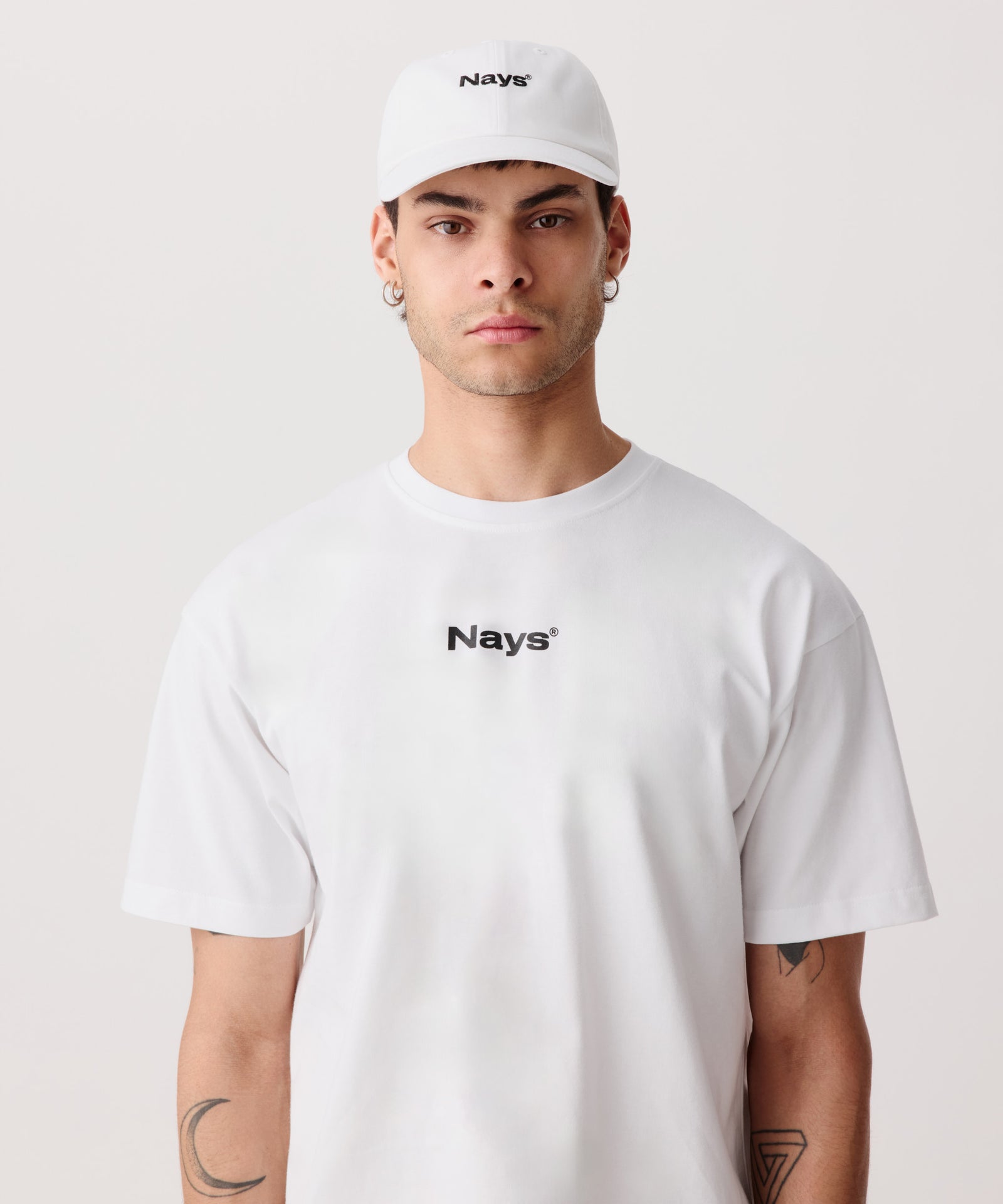 Eine weiße, sportliche Schirm-Mütze mit mittig gesticktem Nays Logo in schwarz. Das Model trägt dazu ein weißes T-Shirt mit einem schwarzen Nays Logo.