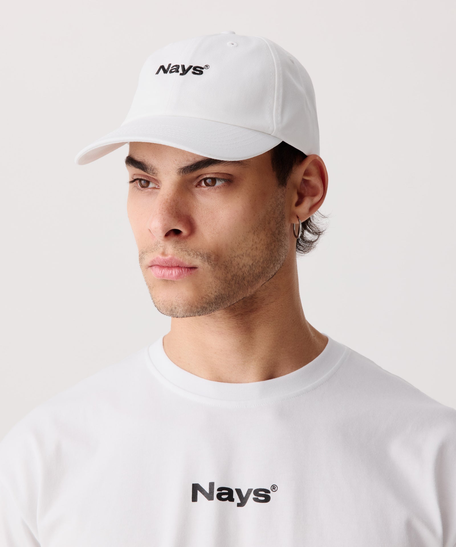 Eine weiße, sportliche Schirm-Mütze mit mittig gesticktem Nays Logo in schwarz. Das Model trägt dazu ein weißes T-Shirt mit einem schwarzen Nays Logo.