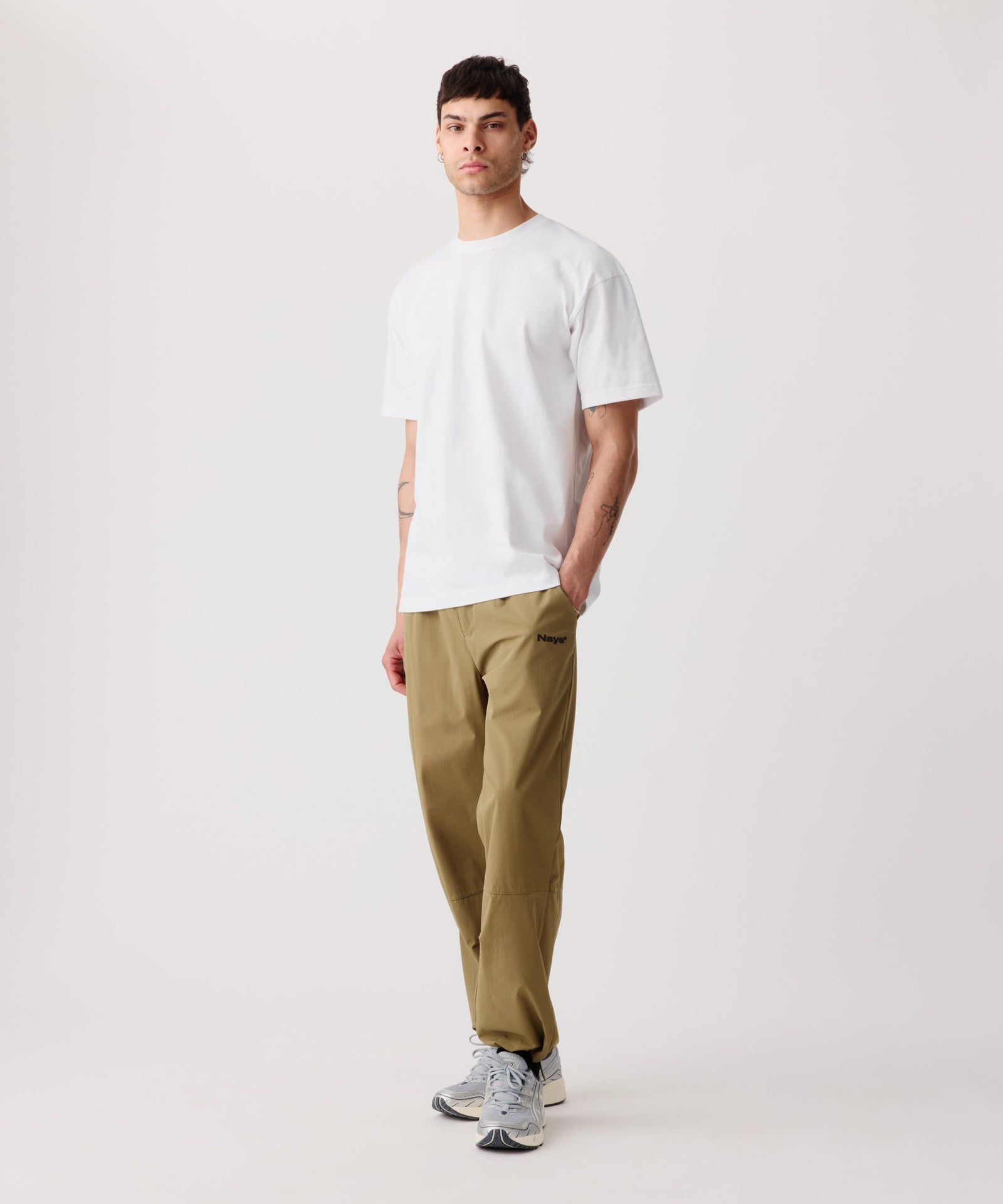 Ein Mann trägt unser Logo T-Shirt mit Rücken-Print. Kombiniert dazu eine beige, lange Hose und helle Turnschuhe. Eine Hand steckt in der Seiten-Tasche.