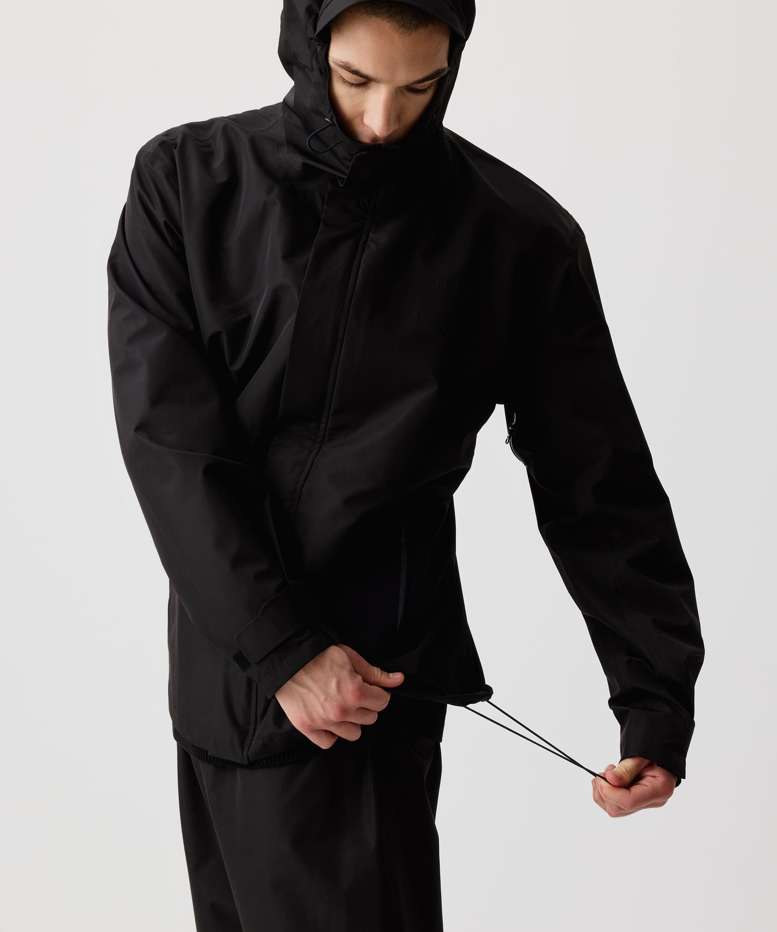 Ein Mann trägt die Nays 3L Multifunctional Rain Jacket mit verstellbarer Kapuze und stellt den Saum der Jacke mit dem elastischen Band ein. YKK AquaGuard® Reißverschlüssen und magnetischer Knopfleiste für optimalen Wetterschutz.