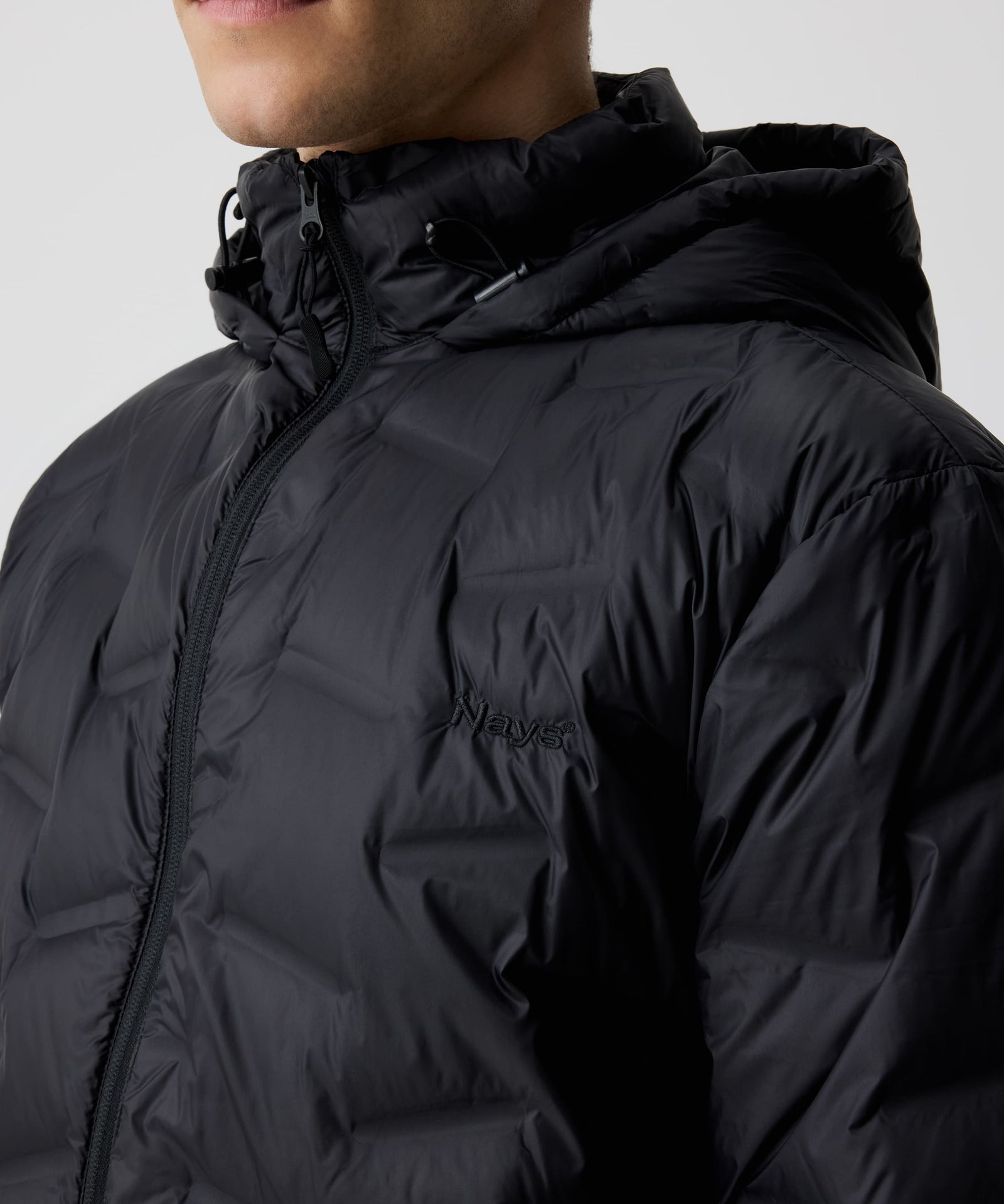 Ein Mann trägt die Nays Lightweight Down Jacket. Der Fokus liegt auf dem YKK-Reißverschluss und dem gestickten, tonalen Logo auf der Brust.
