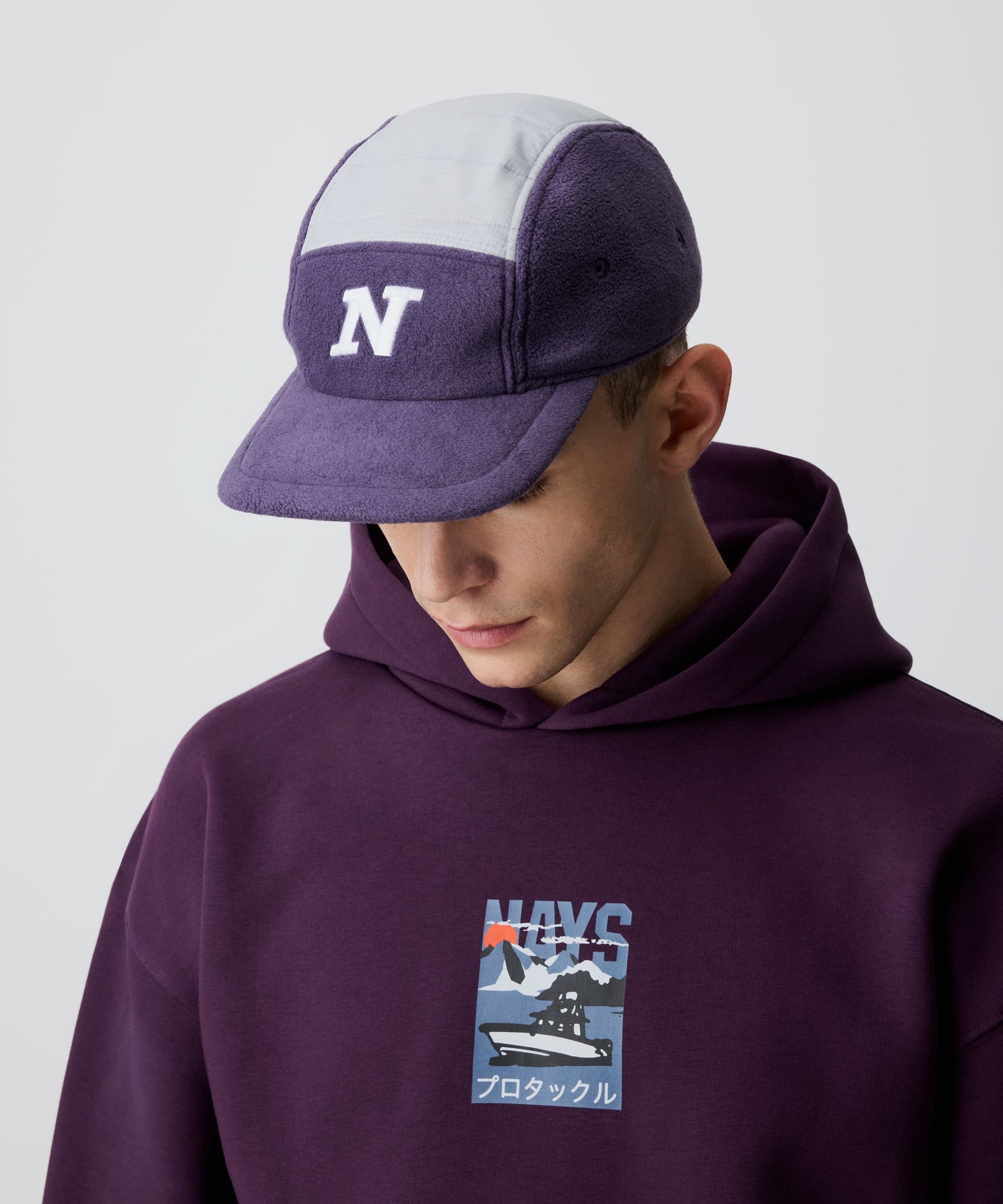 Nays Sherpa 5 Panel Cap mit weißem 'N' vorne, aus weichem flieder-farbigen Sherpa-Material und gefüttertem, weiß abgesetzten Ripstop am Kopf getragen von einem Mann. Er trägt dazu einen bordeaux-farbigen Kapuzen-Pullover.