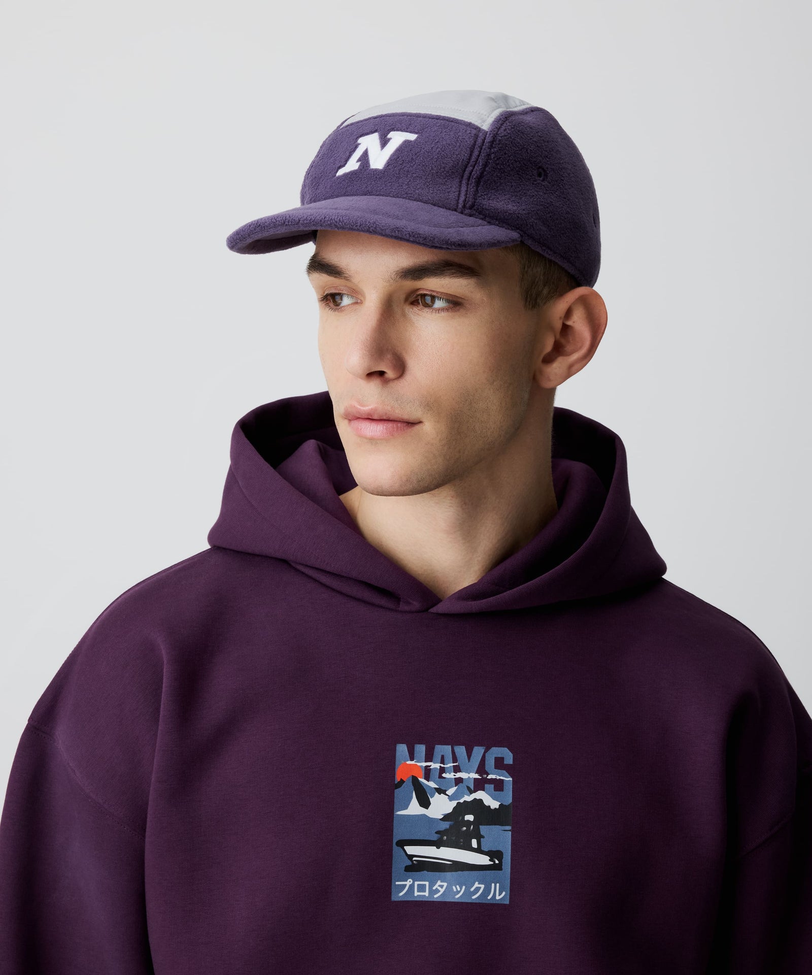 Nays Sherpa 5 Panel Cap mit weißem 'N' vorne, aus weichem flieder-farbigen Sherpa-Material und gefüttertem, weiß abgesetzten Ripstop am Kopf getragen von einem Mann. Er trägt dazu einen bordeaux-farbigen Kapuzen-Pullover mit Print.