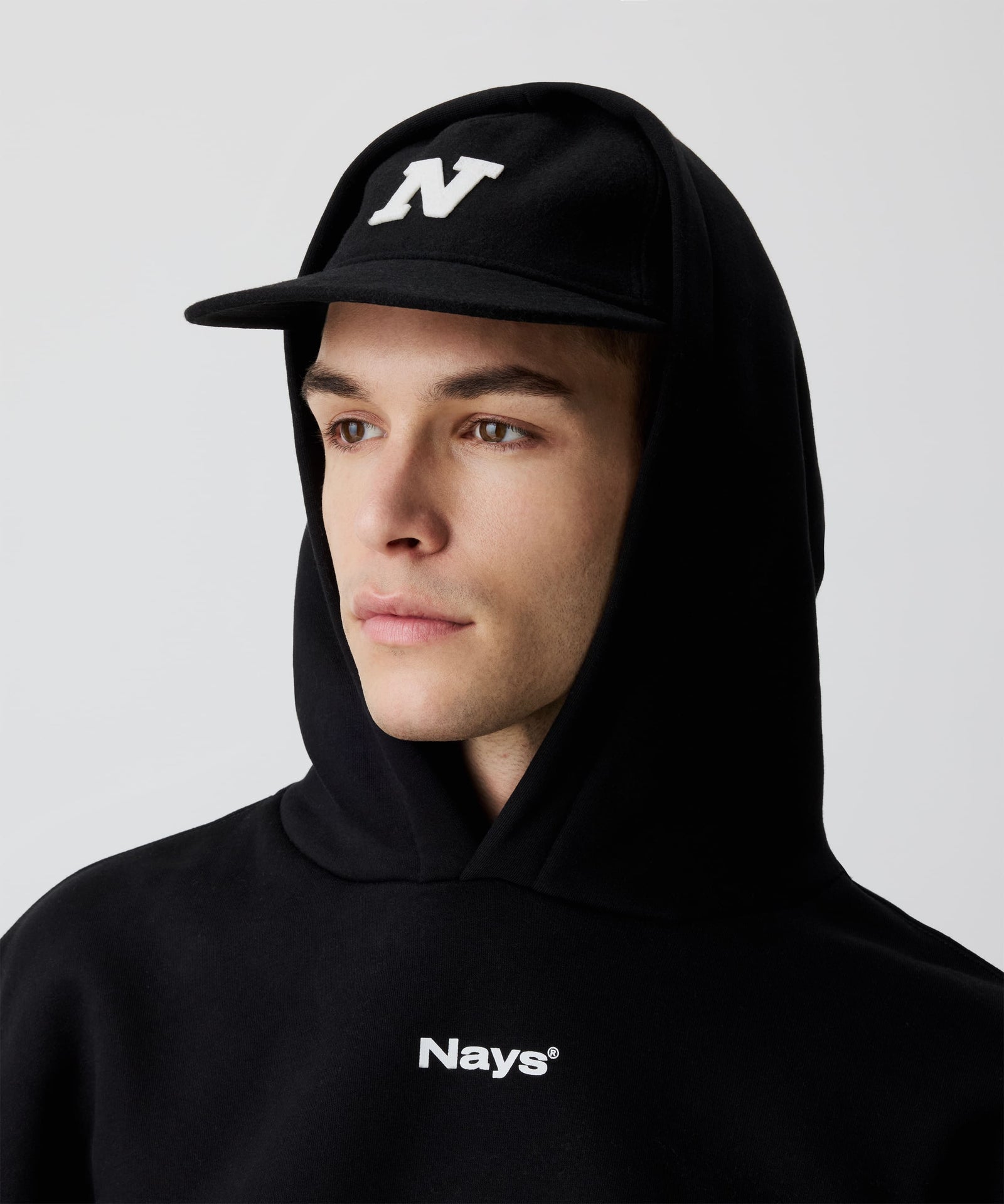 Mann trägt schwarze Nays Felt Cap, eine Schirmmütze aus Filz kombiniert mit einem schwarzen Kapuzen-Pullover mit weißem Nays-Logo-Print auf der Brust.