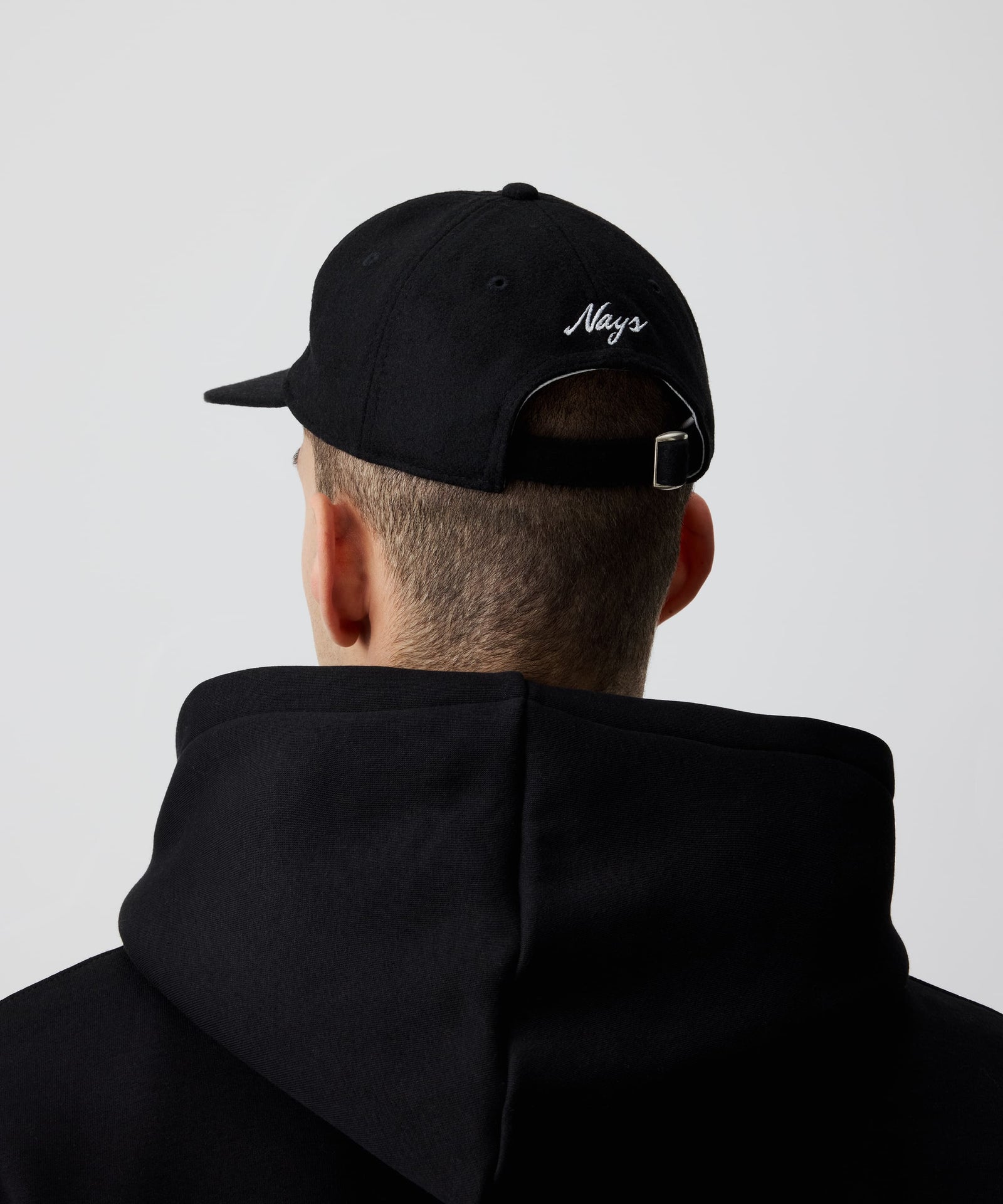 Mann trägt schwarze Nays Felt Cap, eine Schirmmütze aus Filz kombiniert mit einem schwarzen Kapuzen-Pullover in der Rückansicht. Zu erkennen sind das weiße Logo und der größen-verstellbare Riemen.