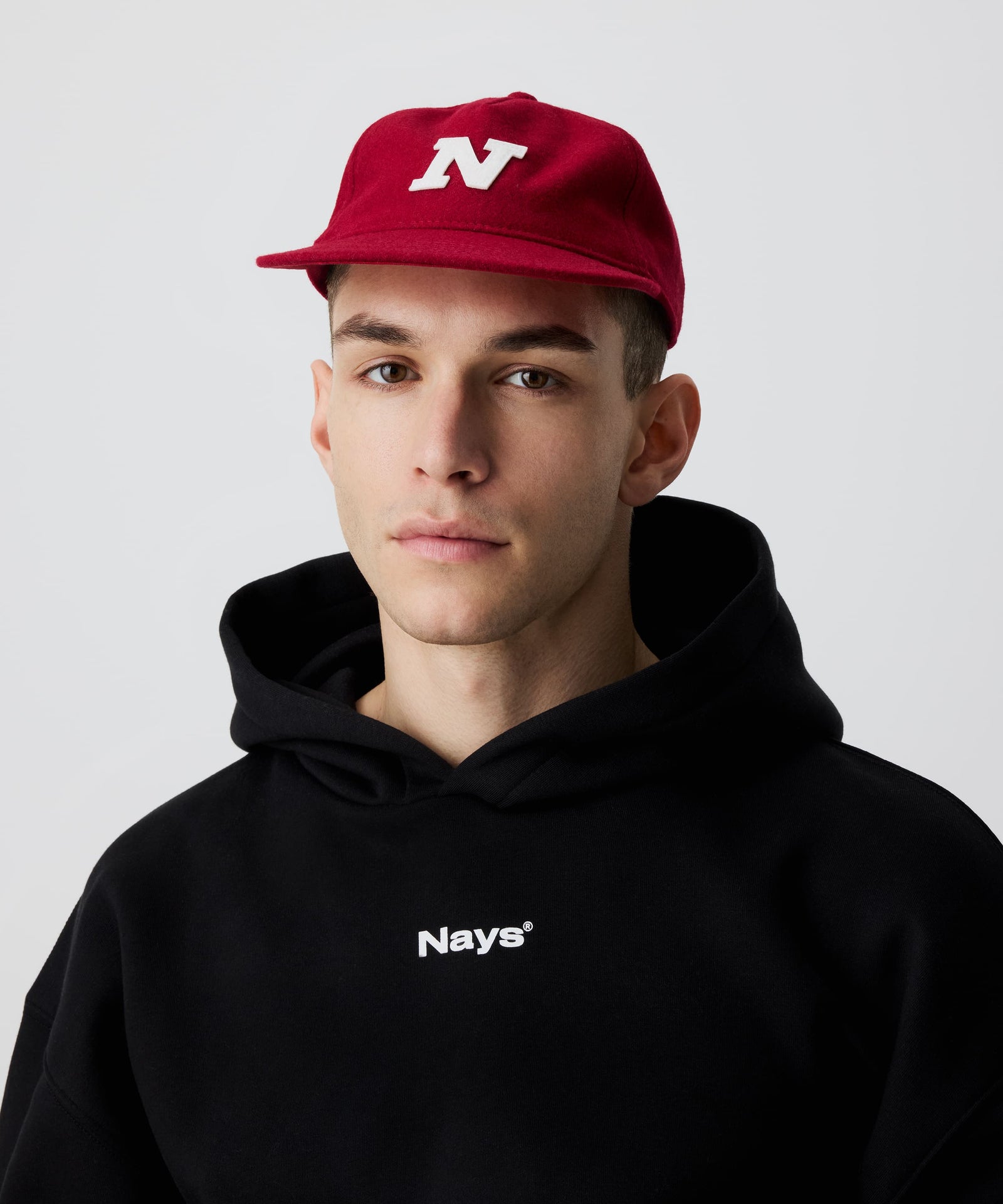 Mann trägt rote Nays Felt Cap, Schirmmütze aus Filz kombiniert mit einem schwarzen Kapuzen-Pullover mit weißem Nays-Logo-Print auf der Brust.