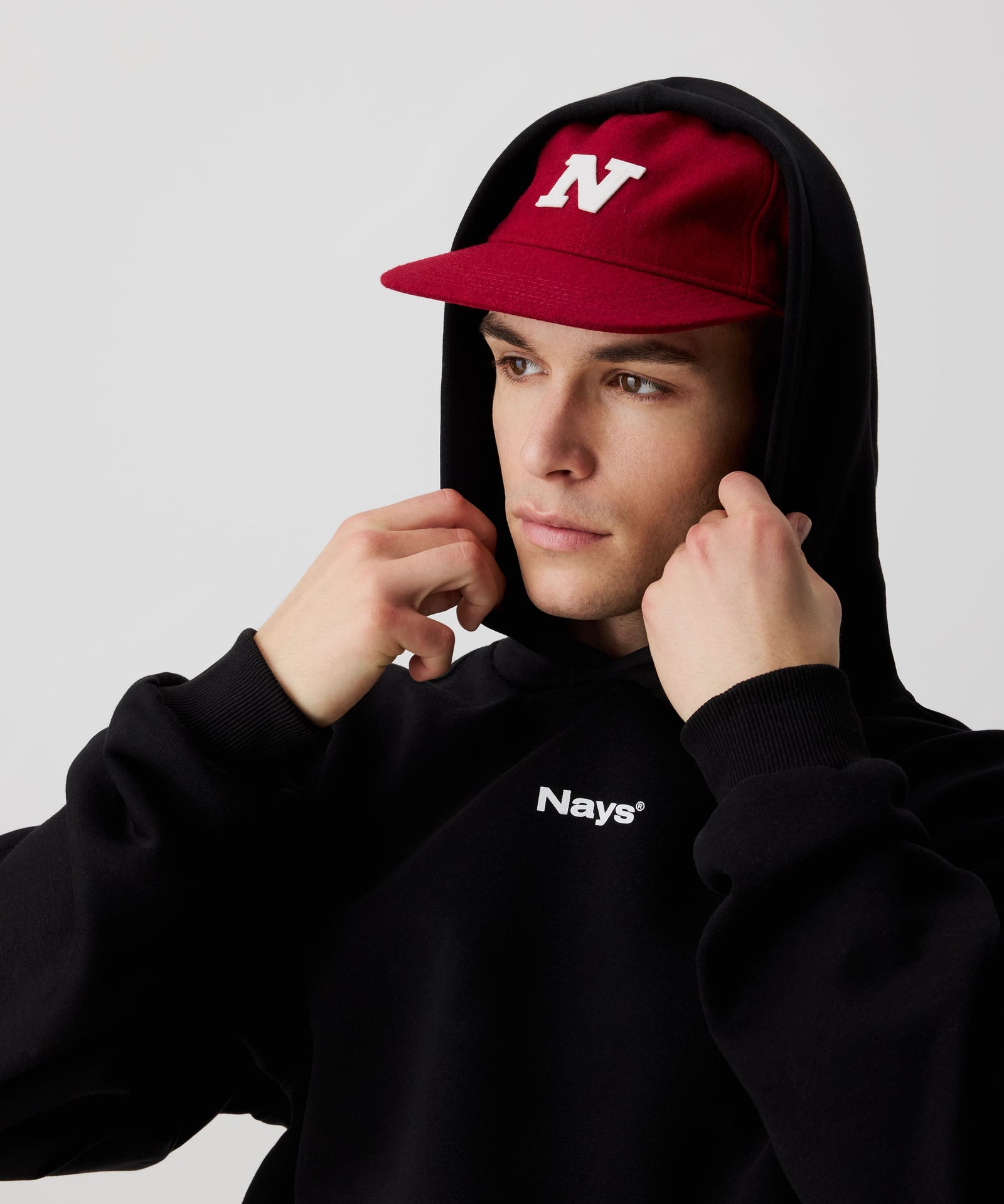 Mann trägt rote Nays Felt Cap, eine stilvolle, robuste Filzmütze kombiniert mit einem schwarzen Kapuzen-Pullover mit weißem Nays-Logo-Print auf der Brust.