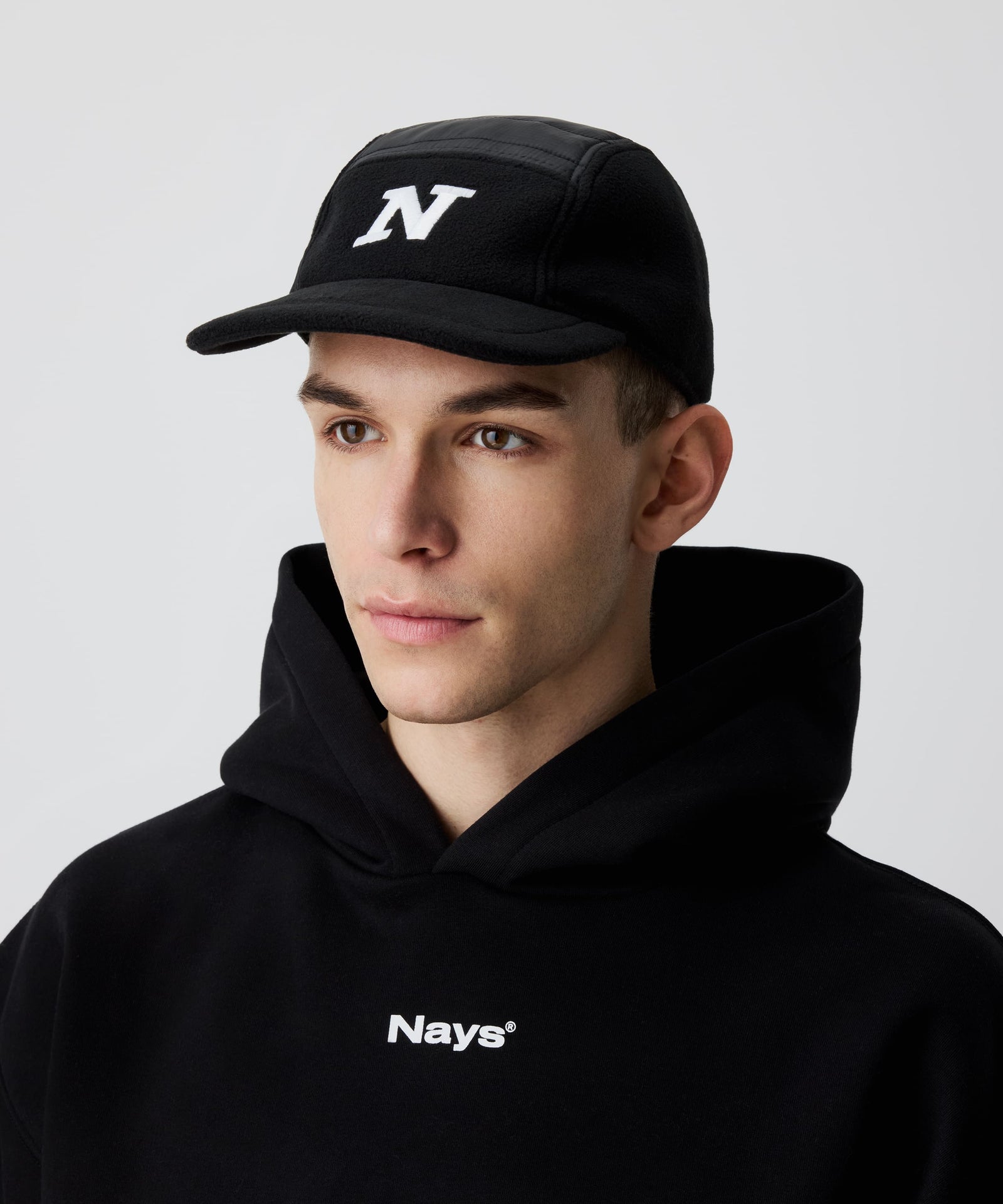 Nays Sherpa 5 Panel Cap mit weißem 'N' vorne, getragen von einem Mann und kombiniert mit einem schwarzen Kapuzen-Pullover mit weißem Logo.