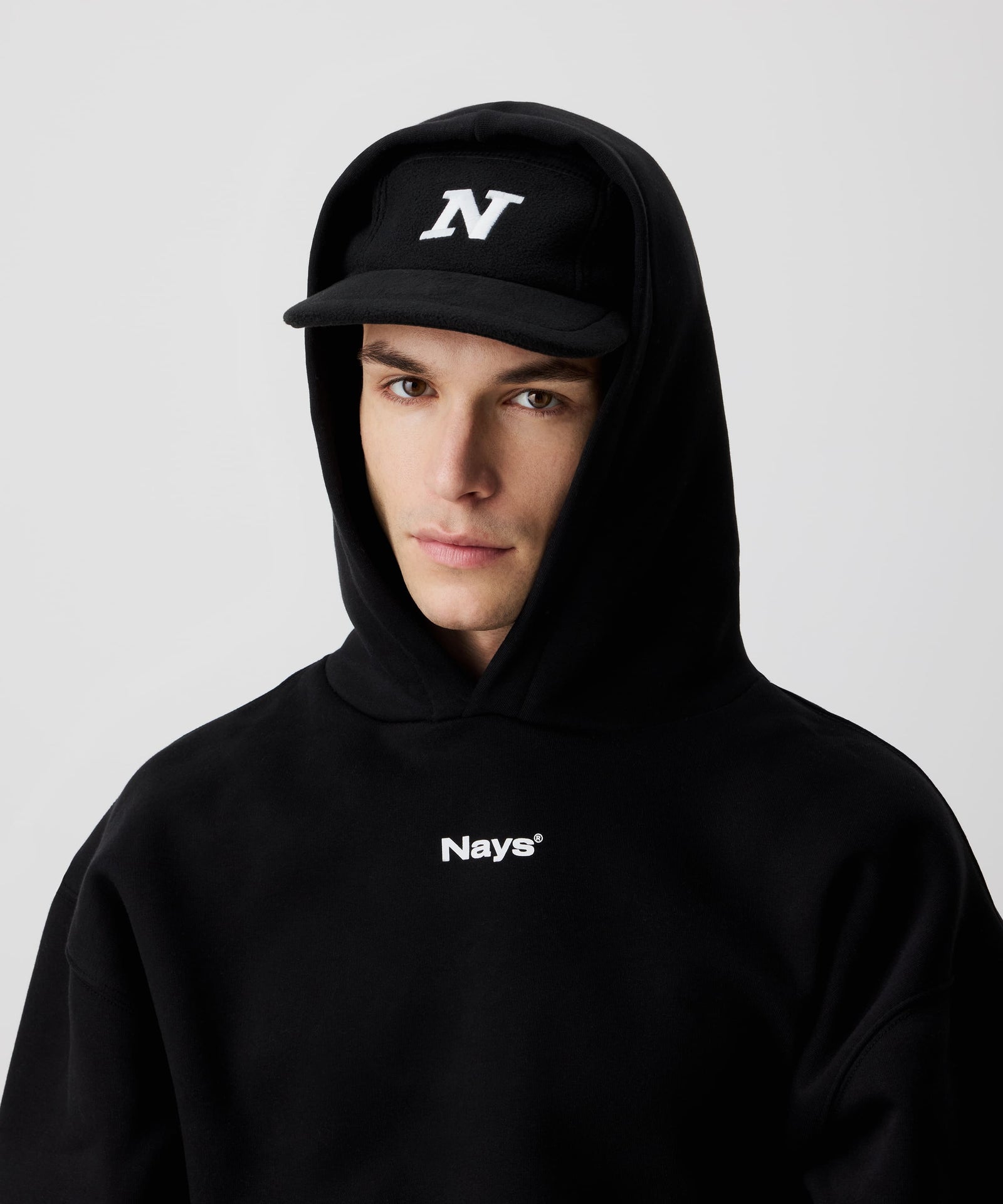 Nays Sherpa 5 Panel Cap mit weißem 'N' vorne, getragen von einem Mann und kombiniert mit einem schwarzen Kapuzen-Pullover mit weißem Logo. Der Mann trägt die Kapuze übern der Mütze auf dem Kopf.