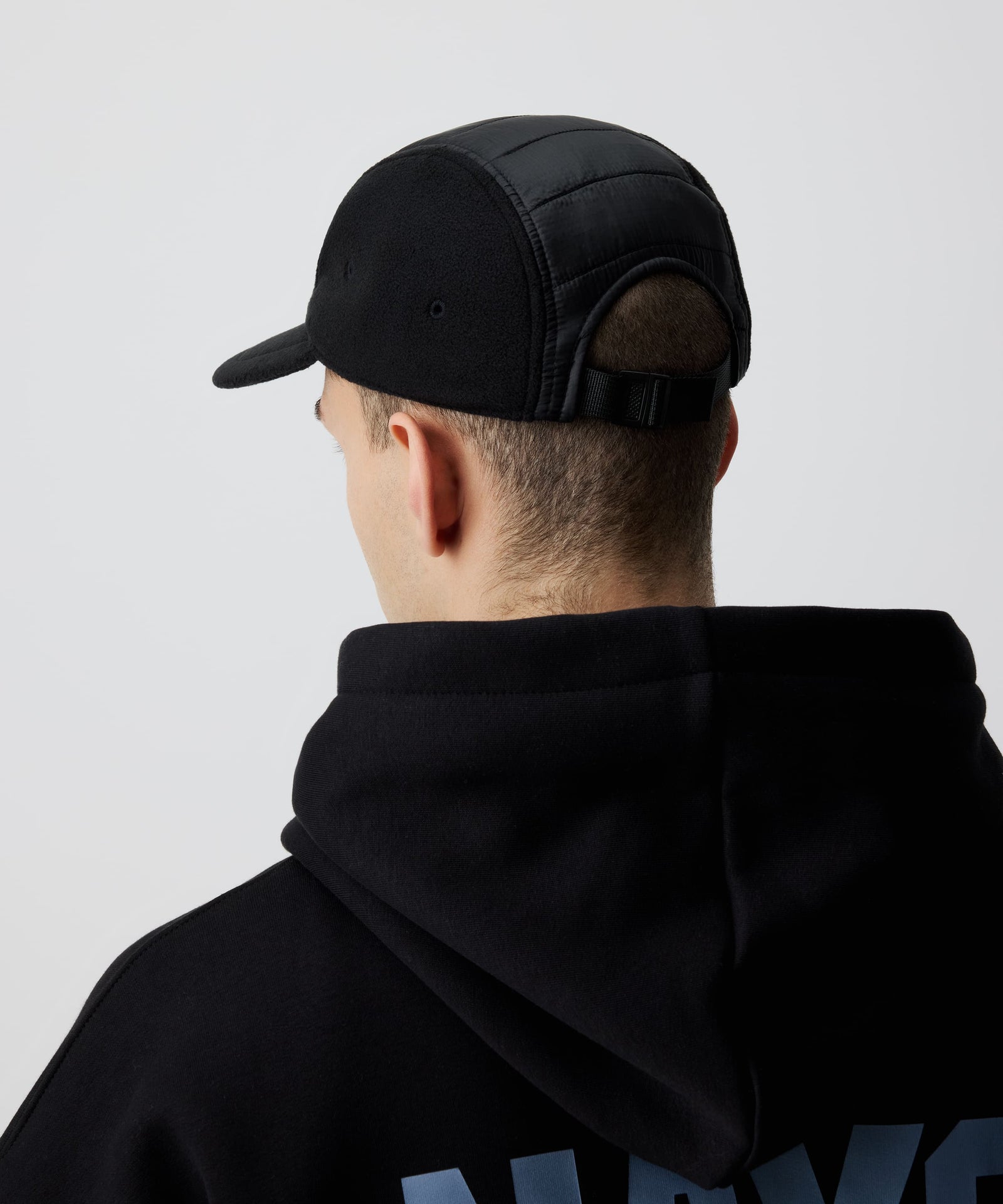 Schwarze Nays Sherpa 5 Panel Cap getragen von einem Mann und kombiniert mit einem schwarzen Kapuzen-Pullover von hinten. Gut zu sehen ist der Material-Mix und der größen-verstellbare Riemen.
