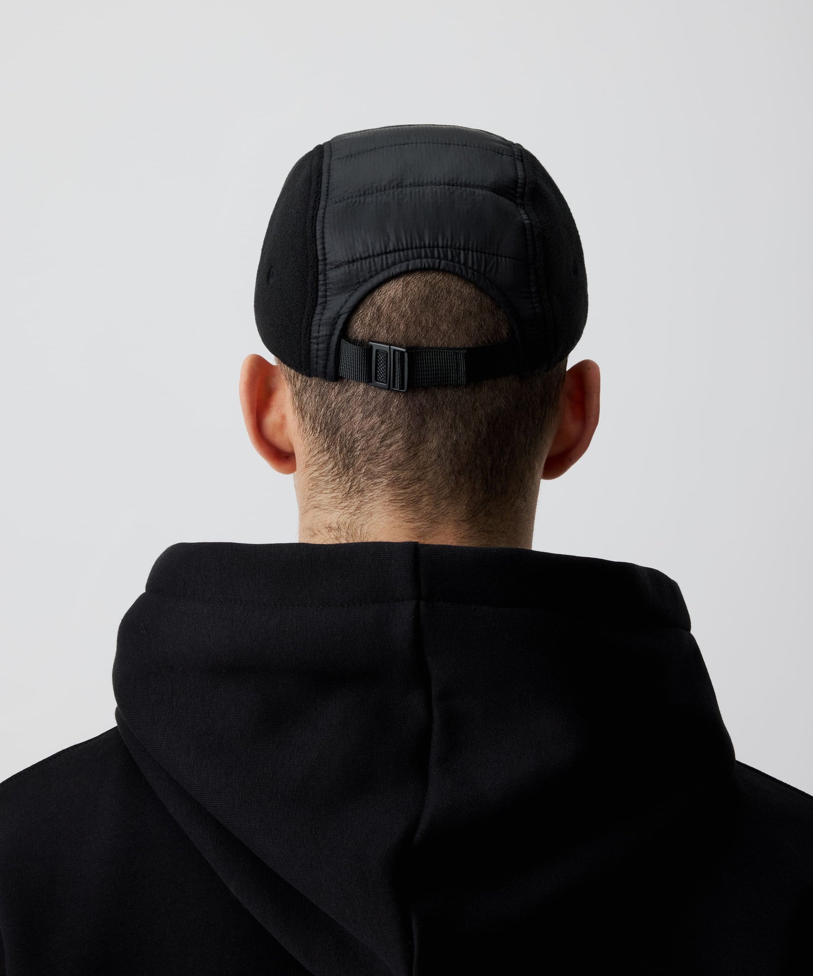 Schwarze Nays Sherpa 5 Panel Cap getragen von einem Mann und kombiniert mit einem schwarzen Kapuzen-Pullover von hinten. Gut zu sehen ist der Material-Mix und der größen-verstellbare Riemen.