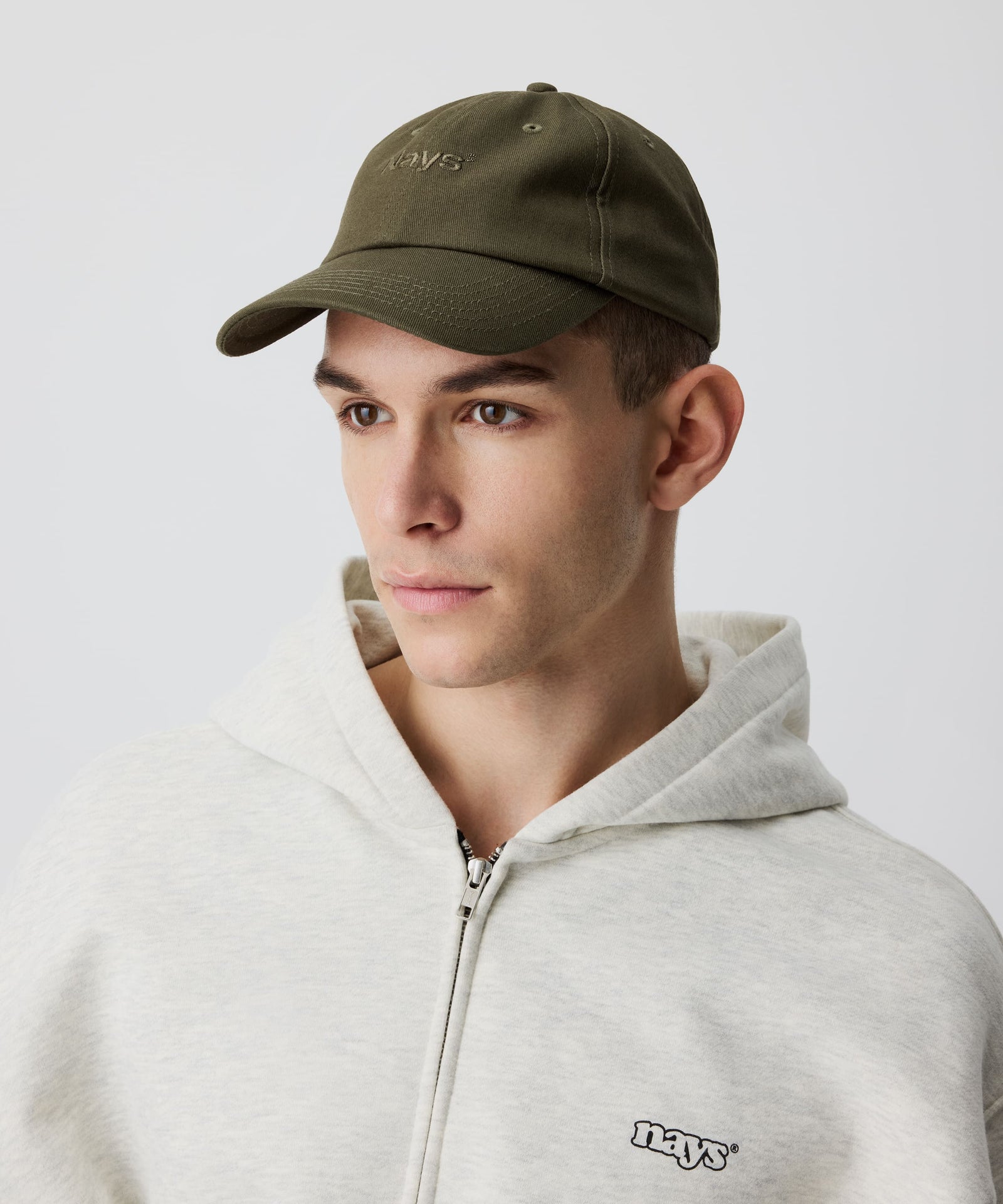 Das Detail-Bild zeigt unsere khaki-farbene Schirm-Mütze getragen von einem Mann. Er trägt dazu einen hell-grauen Kapuzen-Pullover mit Reißverschluss.