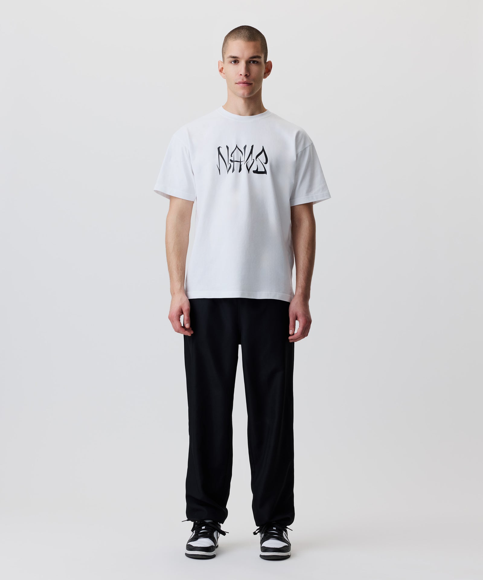 Das Bild zeigt die schwarze Hose getragen von einem Mann. Er trägt dazu ein weißes T-Shirt mit schwarzem Logo und schwarz-weiße Turnschuhe.
