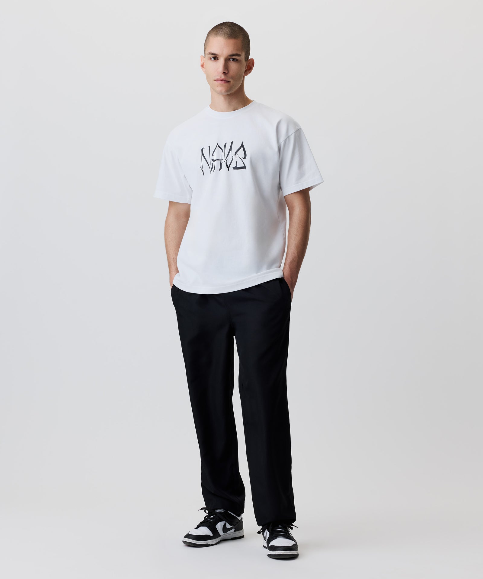 Das Bild zeigt die schwarze Hose getragen von einem Mann. Er trägt dazu ein weißes T-Shirt mit schwarzem Logo und schwarz-weiße Turnschuhe. Beide Hände stecken in den Hosentaschen.