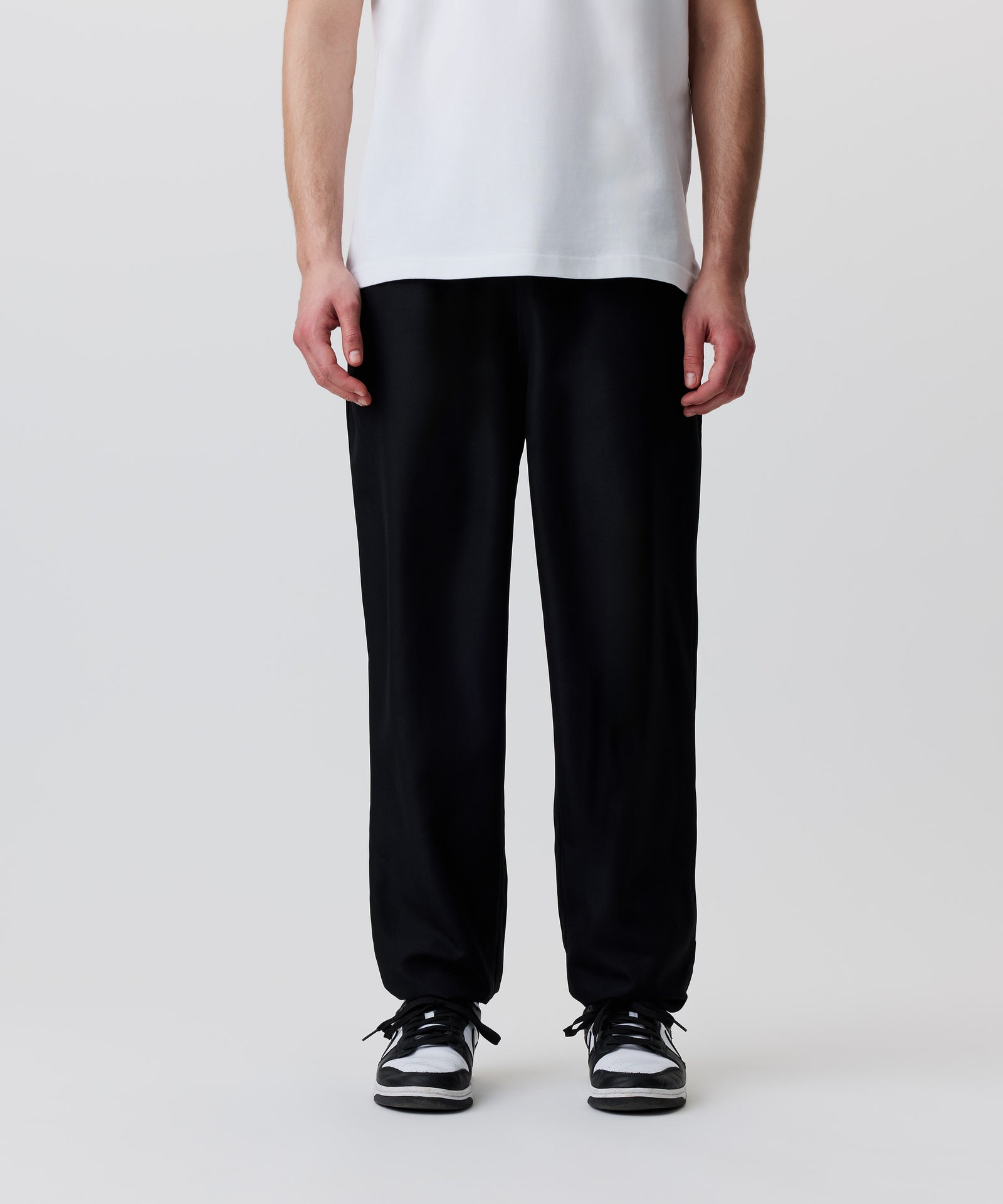 Der Bild-Ausschnitt zeigt die schwarze Hose getragen von einem Mann. Er trägt dazu ein weißes T-Shirt und schwarz-weiße Turnschuhe.