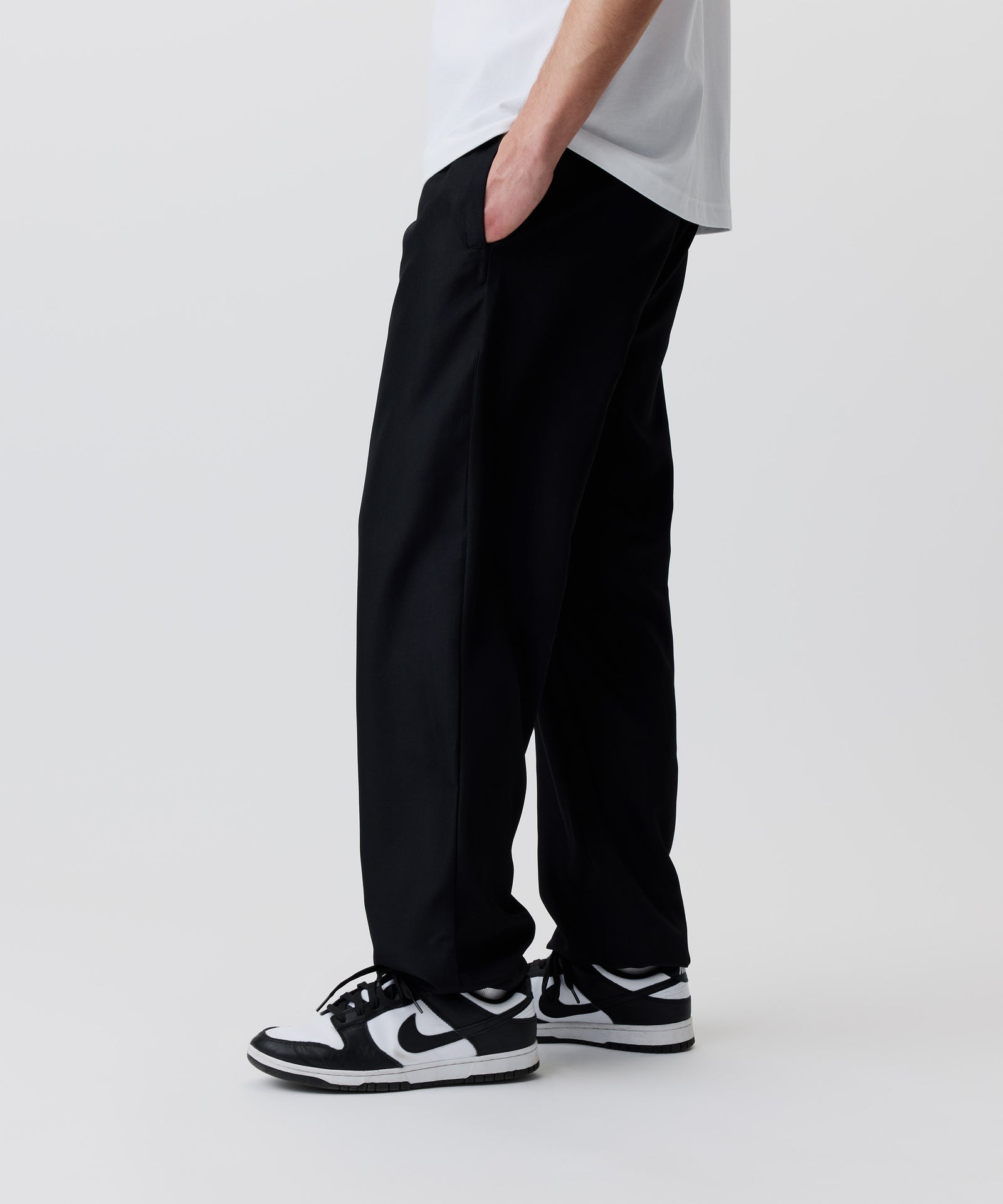 Der Bild-Ausschnitt zeigt die schwarze Hose getragen von einem Mann im Seiten-Profil. Er trägt dazu ein weißes T-Shirt und schwarz-weiße Turnschuhe.
