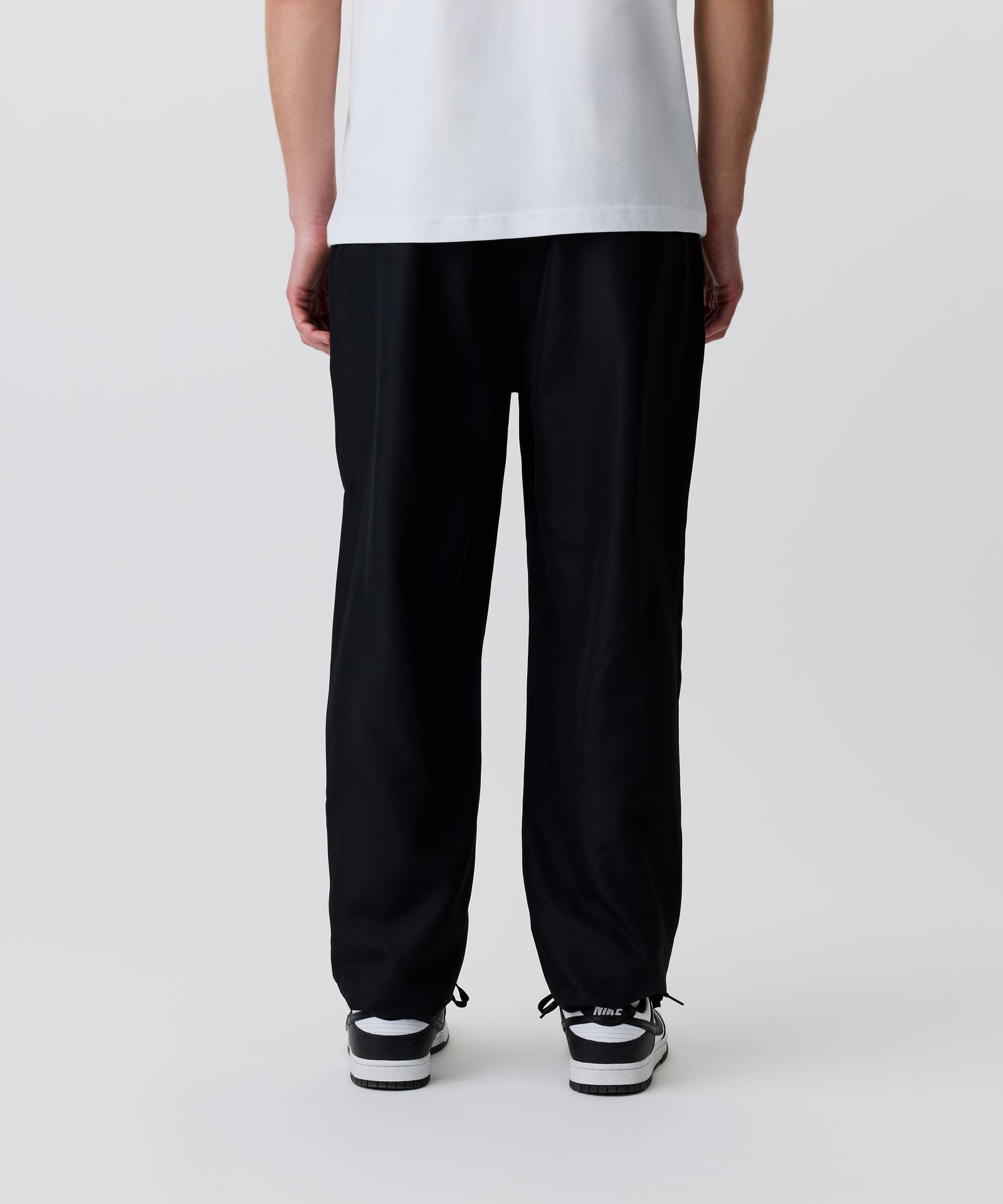 Der Bild-Ausschnitt zeigt die schwarze Hose getragen von einem Mann in der Rückansicht. Er trägt dazu ein weißes T-Shirt und schwarz-weiße Turnschuhe.
