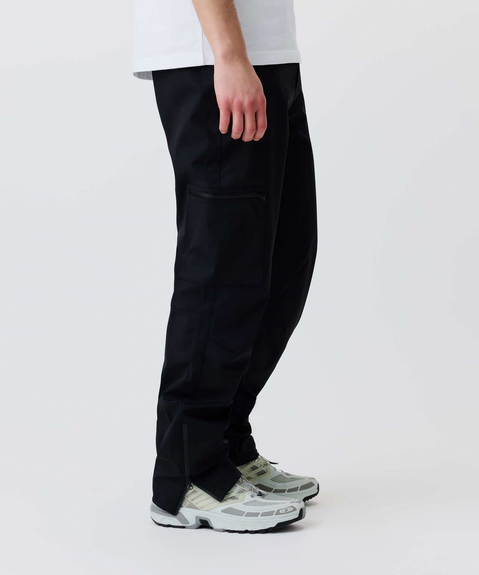 In diesem Bild-Ausschnitt sieht man unsere schwarze Hose getragen von einem Mann im Seiten-Profil. Gut zu erkennen ist ein Reißverschluss am Hosen-Bund. Dazu trägt er helle Turn-Schuhe.