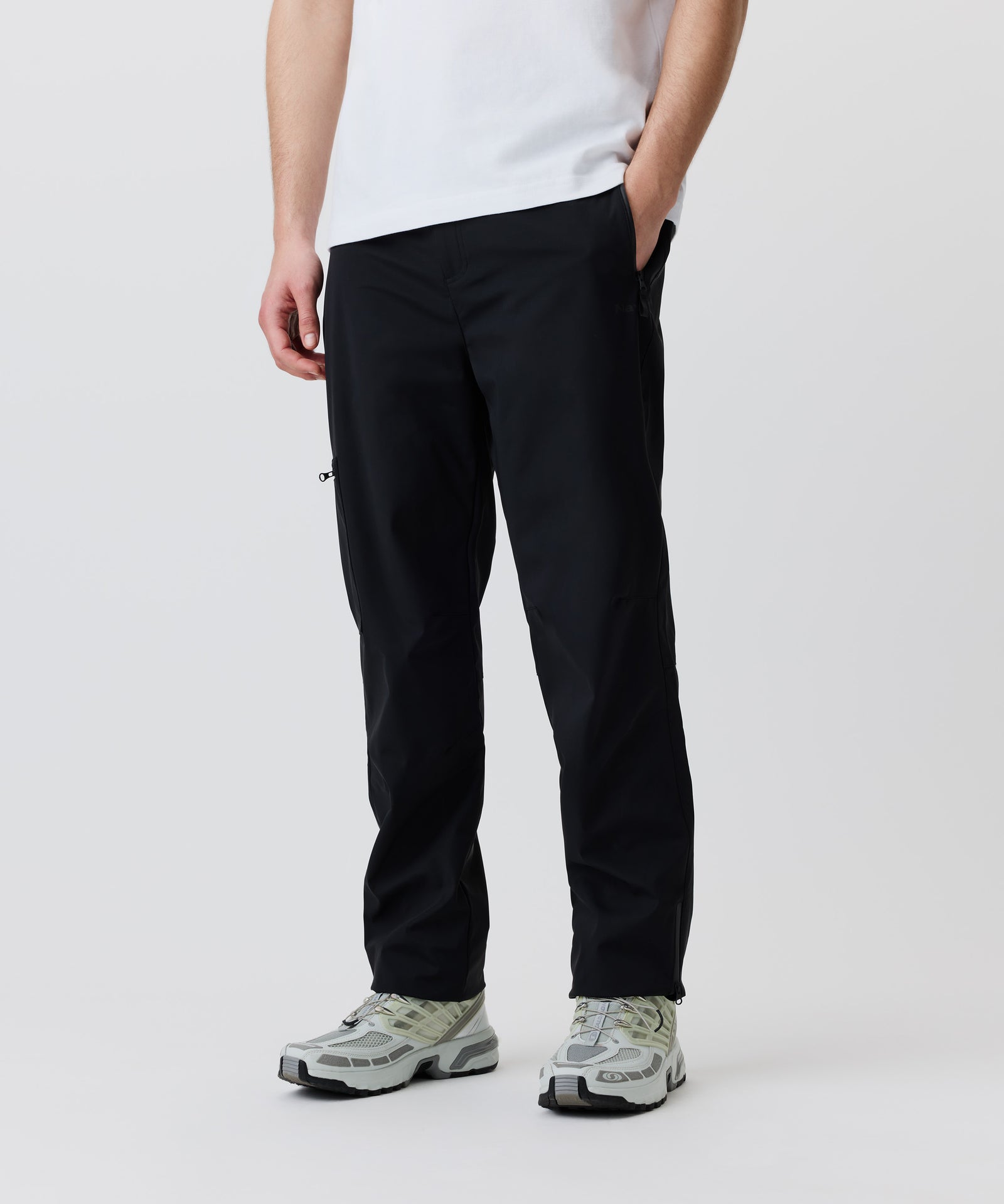In diesem Bild-Ausschnitt sieht man unsere schwarze Hose getragen von einem Mann. Er trägt ein weißes T-Shirt und helle Turn-Schuhe. Seine linke Hand steckt in der Hosen-Tasche.