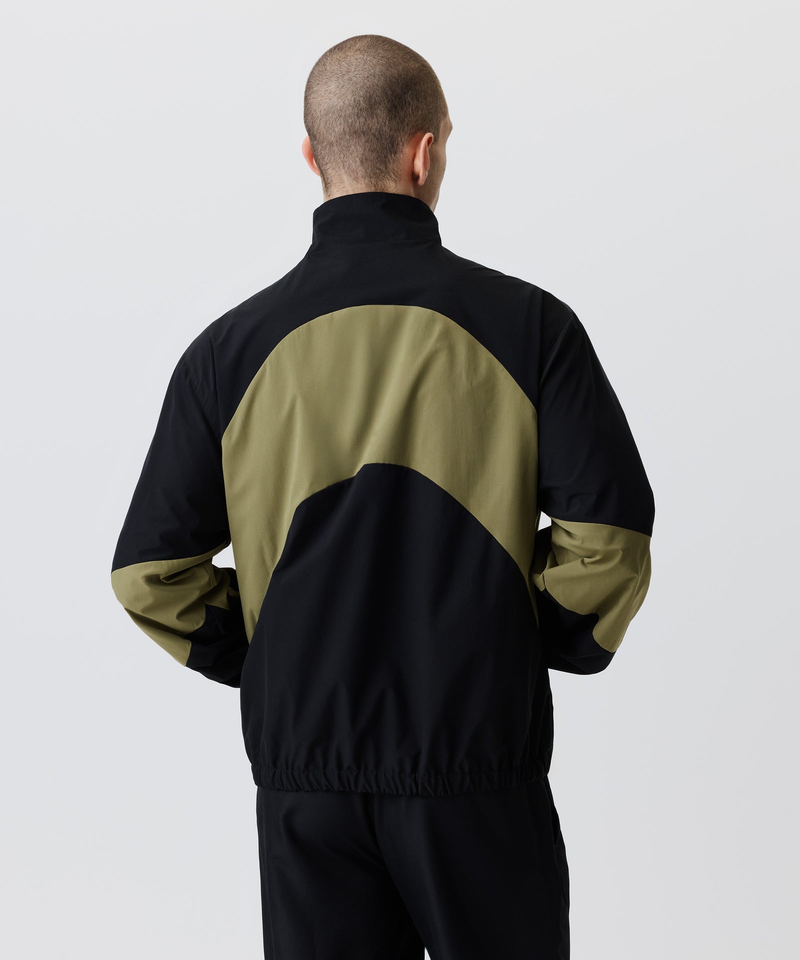 Auf dem Bild in der Rückansicht trägt der Mann den Wind-Breaker in Kombination mit einer schwarzen Hose. beide Hände stecken in der Brust-Tasche.