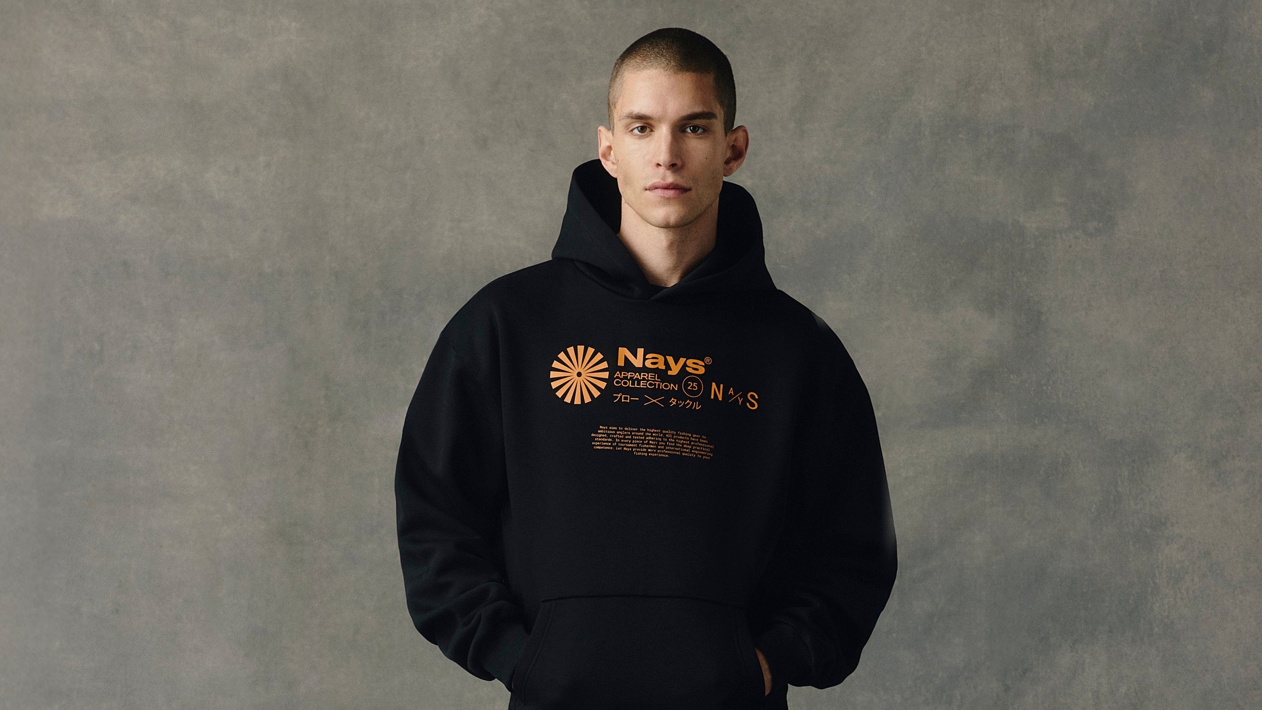 Frontansicht Mann im schwarzen Kapuzen-Pullover mit orange-farbigem Nays Schriftzug vor einem grauem Hintergrund.