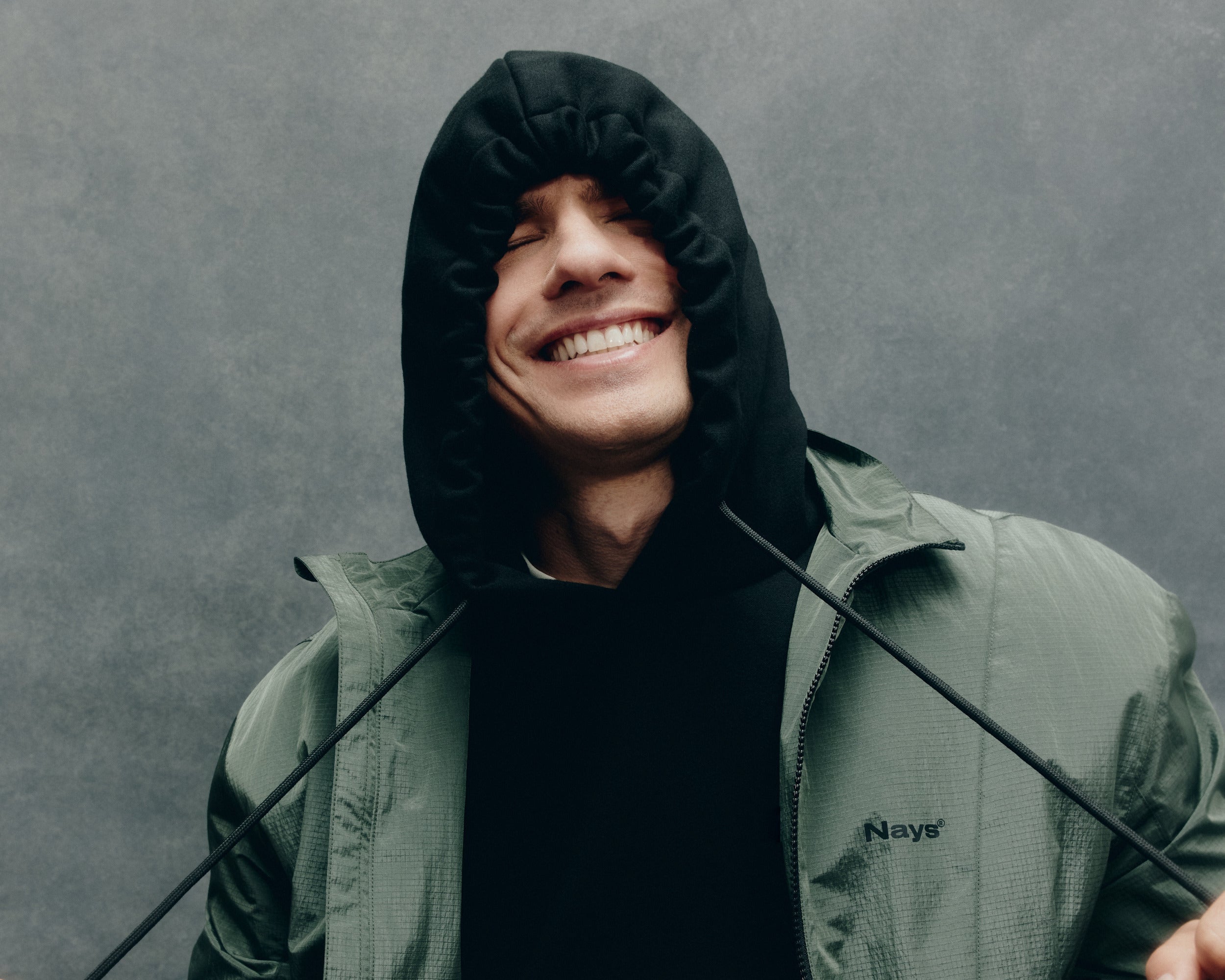 Frontansicht Mann in grüner Jacke vor einem grauem Hintergrund. Die Kapuze ist aufgesetzt und er zieht sie an den Kordeln tief ins Gesicht.