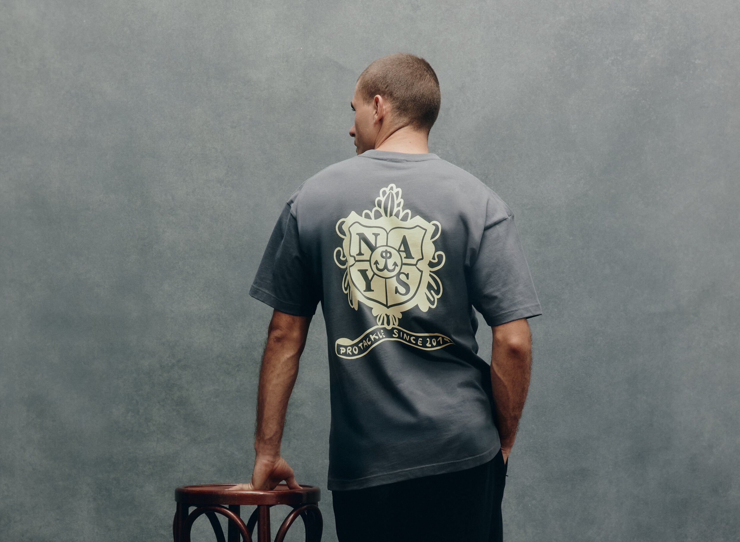 Rückansicht Mann im grauen T-Shirt mit hellem Nays-Emblem-Logo-Druck vor einem grauem Hintergrund.