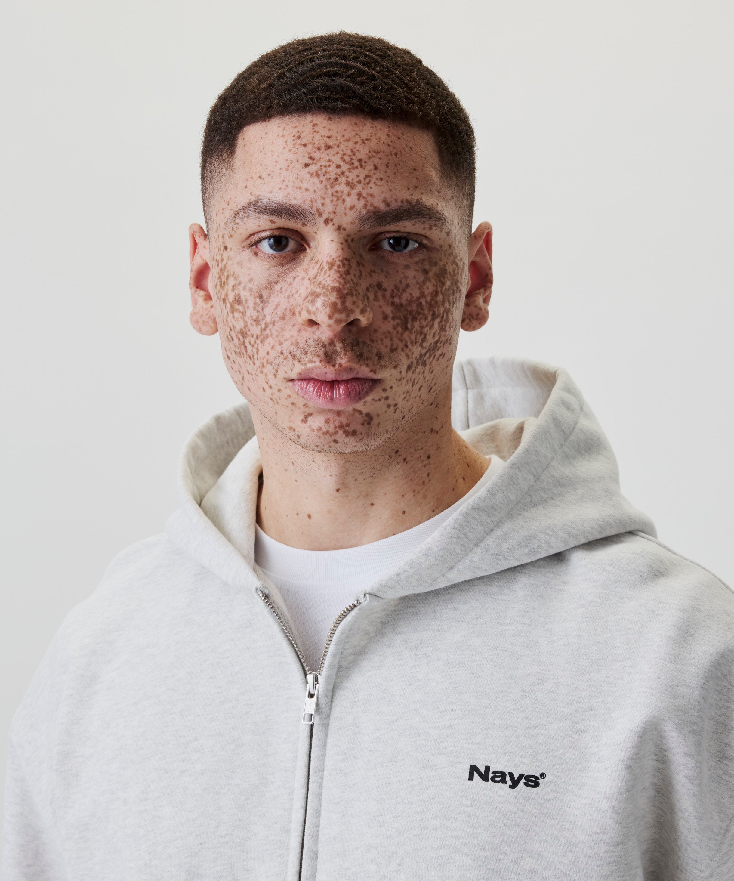Detailaufnahme eines hellgrauen Kapuzenpullovers mit Reißverschluss und schwarzem „Nays“-Logostick auf der Brust.