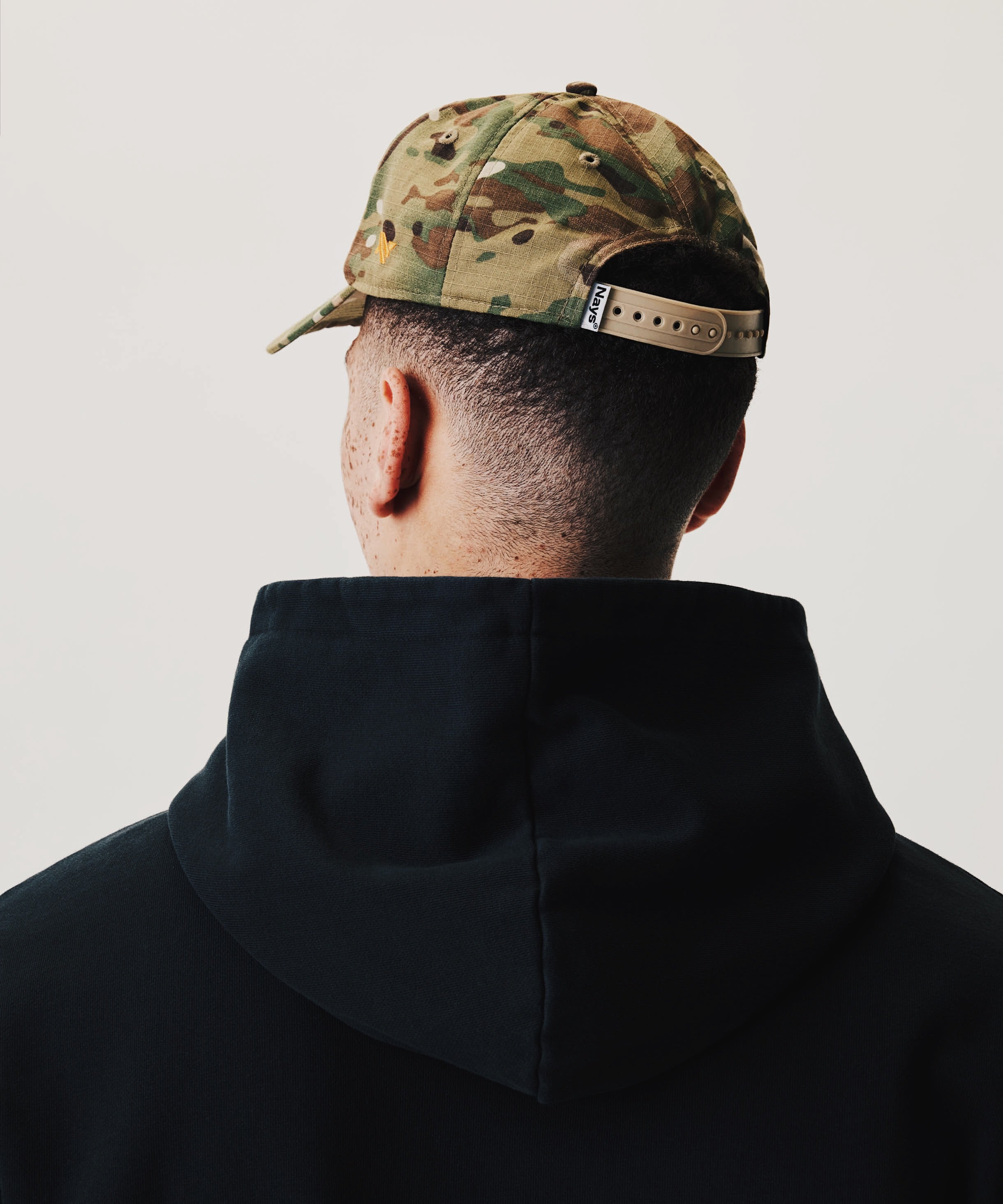 Rückansicht eines Models mit camouflagefarbener NAYS Fishing Club Cap und schwarzem NAYS new Script Hoodie vor hellem Studiohintergrund.