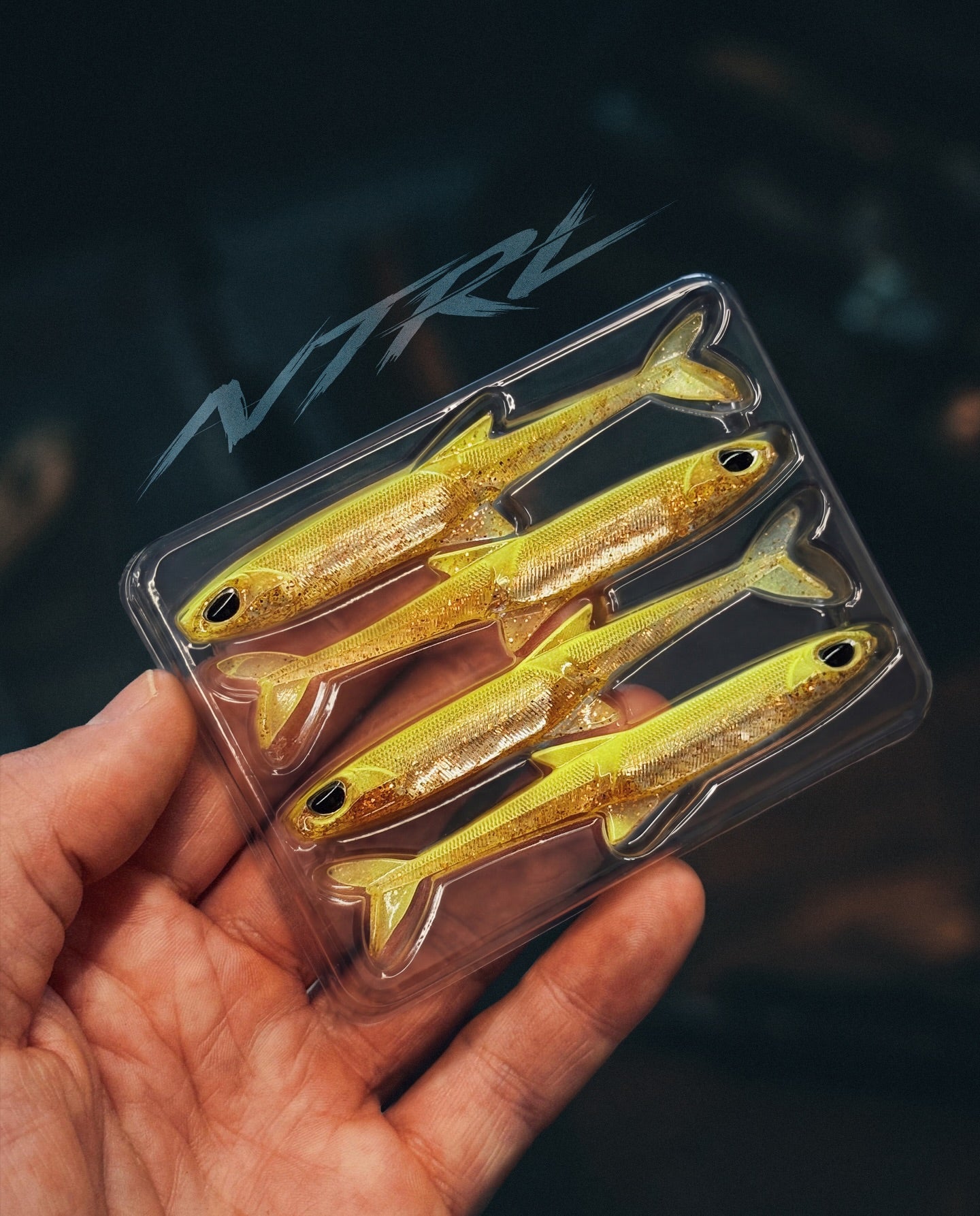 Hand hält eine Blisterverpackung mit gelb-glitzernden Gummifischen der Marke NTRL zum Raubfischangeln.