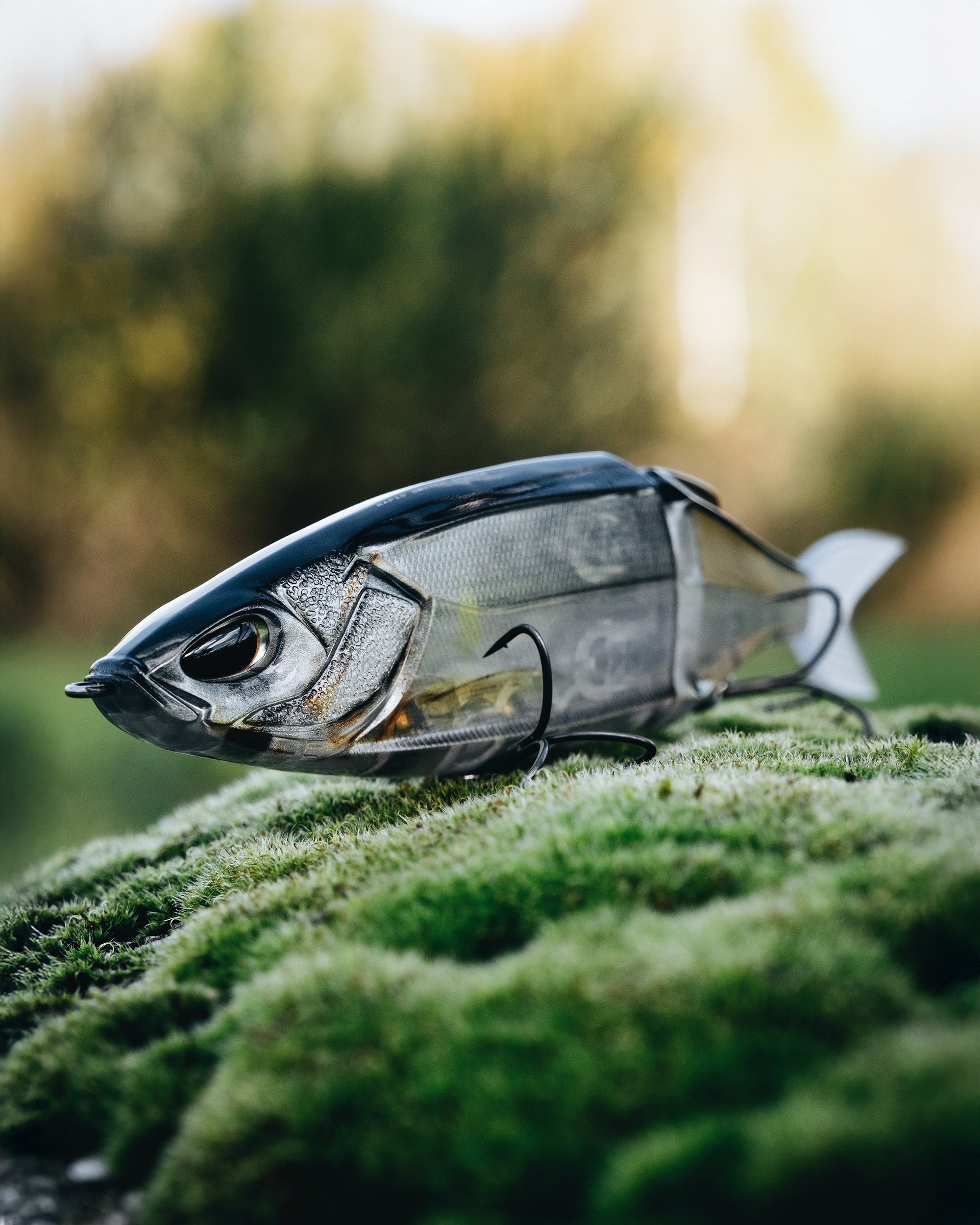 Nahaufnahme eines zweiteiligen, semi-transparenten Angelköder mit einem realistisch detaillierten, silbernen Kopf und zwei Drillings-Haken. Der Köder liegt auf grünem Moos im Freien.