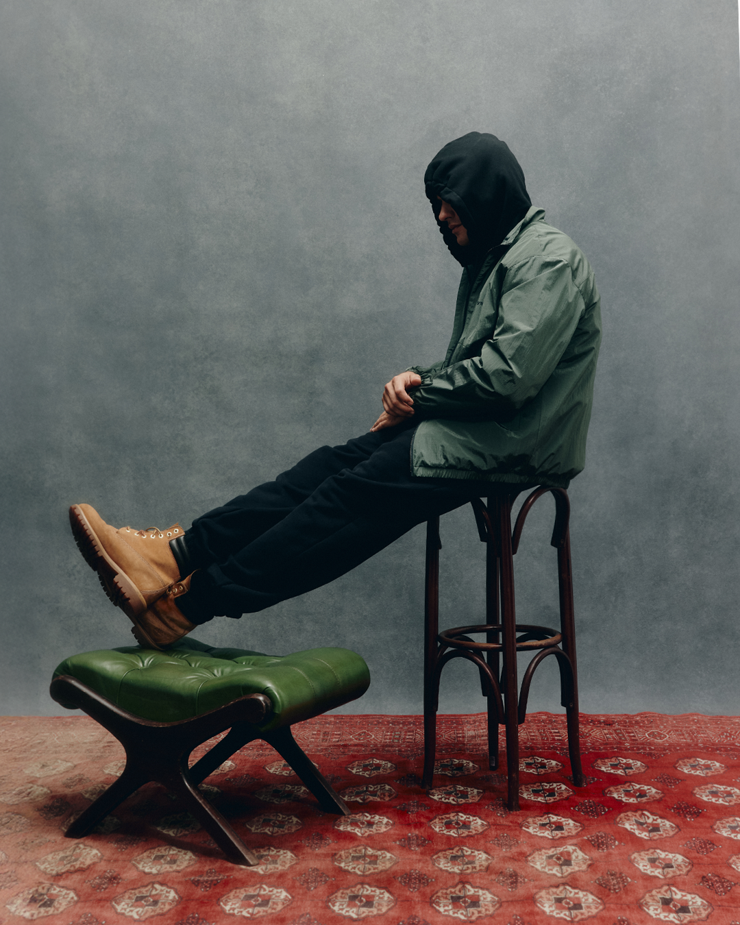 Ein Mann sitzt in grüner Jacke, schwarzer Hose und zugezogener Kapuze auf einem Hocker vor grauem Hintergrund. Die Beine sind auf einem grünen Polsterhocker abgestützt.