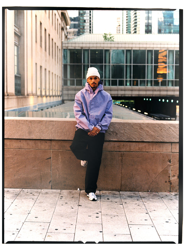 Model in lilafarbener NAYS Rain Jacket 2.0, weißer Beanie und schwarzer Hose vor moderner Stadtarchitektur in Toronto.